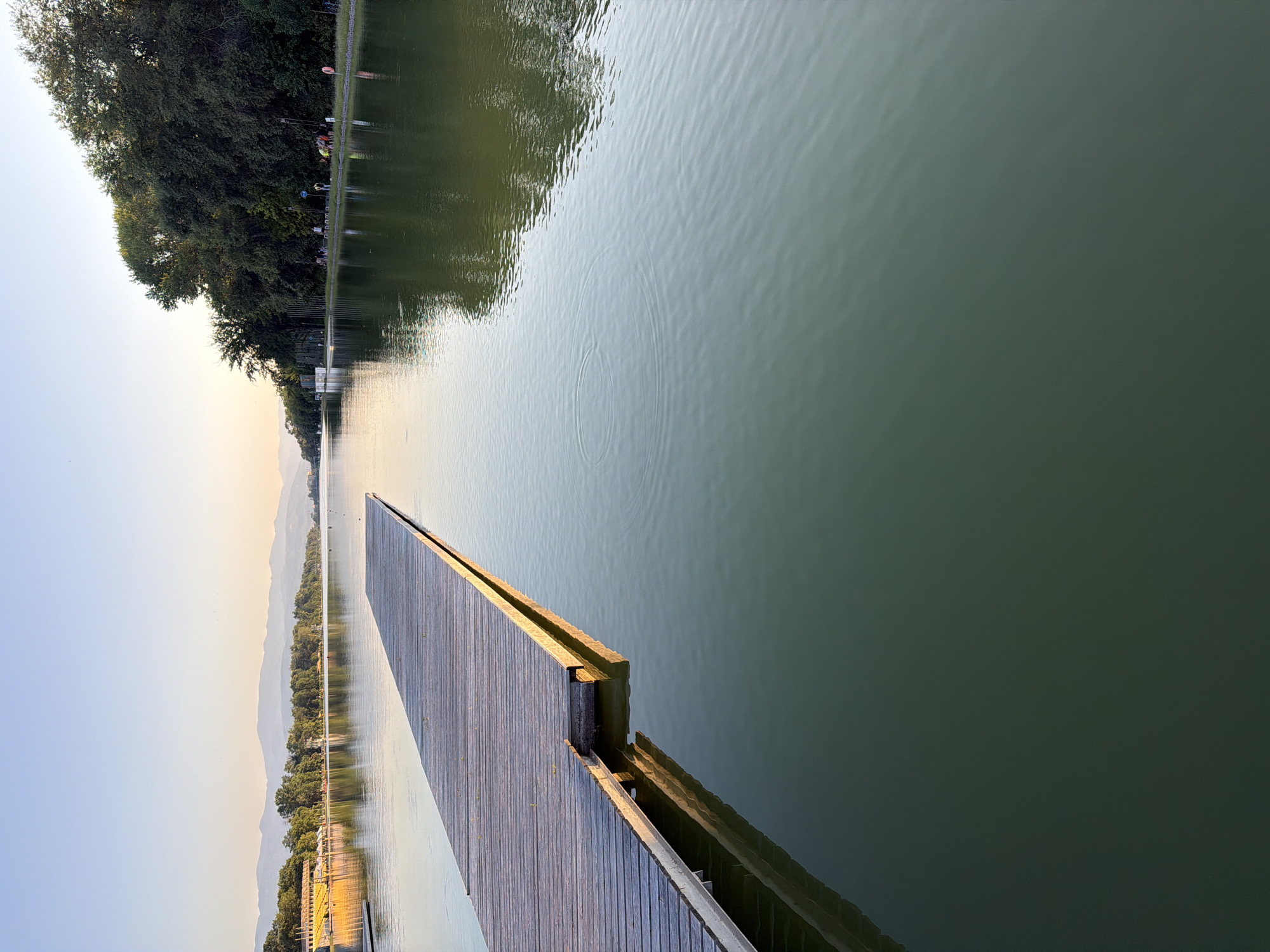 The image captures a serene scene at the Rowing Canal in Plovdiv, Bulgaria, during a clear day with the sun setting on the horizon. A wooden pier extends from the left foreground into the calm, greenish water of the canal, casting a reflection that mirrors its structure. The water surface shows subtle ripples, indicative of a gentle breeze.
In the background, the far bank of the canal is lined with lush green trees, forming a dense forest that gradually recedes towards distant, hazy mountains. On the left side of the canal, a yellow-roofed building, possibly a sports facility or a pavilion, is visible, reflecting in the water. Further along the bank, a few individuals can be seen walking and engaging in leisure activities, their forms softened by the distance. The sky above is a pale blue, transitioning to a soft orange hue near the horizon, suggesting the warm light of dusk. The overall atmosphere is tranquil and picturesque.