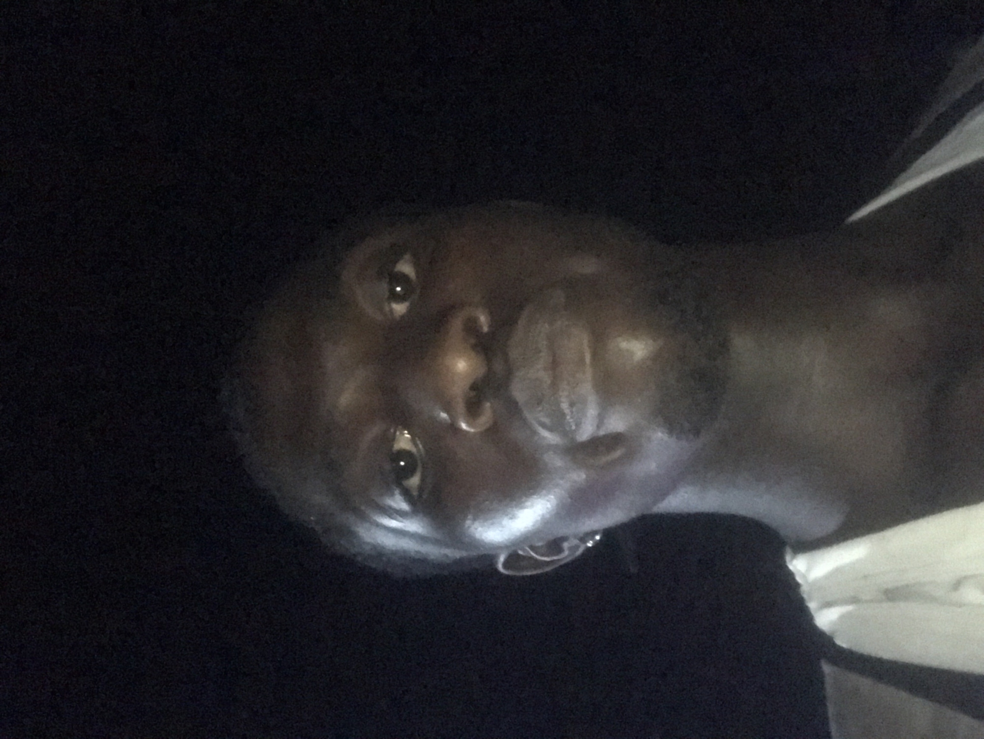 The image presents a portrait of a person, likely male, with dark skin, captured from the chest up. The person's head is rotated approximately 90 degrees counter-clockwise relative to the frame, looking directly towards the camera.
The scene is set against an almost entirely dark background, strongly suggesting a low-light environment or nighttime photography. A direct light source, possibly a camera flash, illuminates the person's face from the front, creating bright highlights on the forehead, nose, and glossy lips. One eye is clearly visible, reflecting the light source, and the person's expression appears neutral or thoughtful.
A small, circular earring is visible on the person's left earlobe. On the right side of the frame, a portion of a garment with white and dark stripes is visible, likely a shirt or top worn by the subject. The overall mood is intimate and focused on the subject due to the stark contrast between the illuminated face and the deep black background. No specific location cues, weather conditions, or visible text are present in the image.