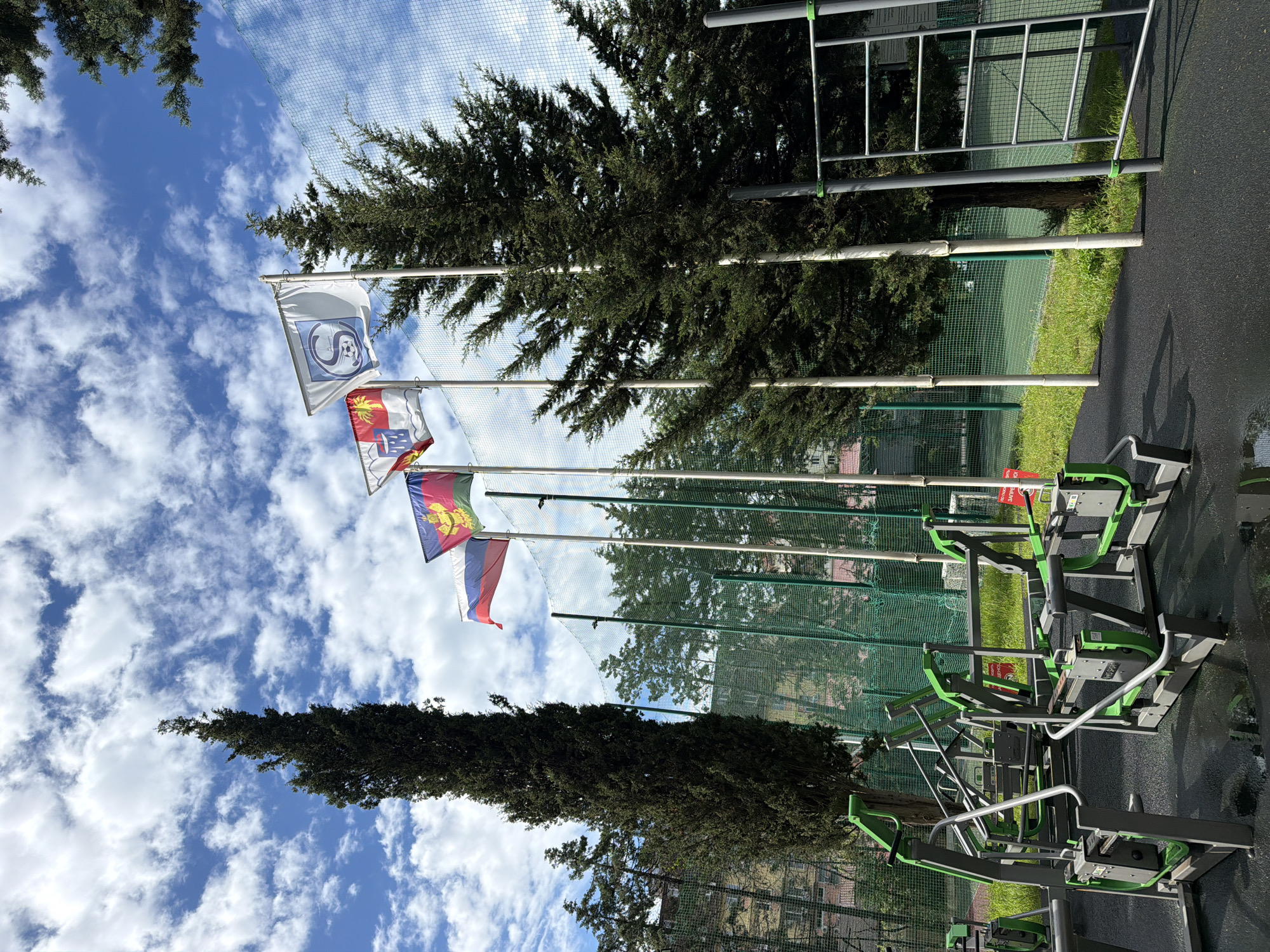 The image shows a view in Sochi, Russia, featuring several flags flying from poles, likely in a park or recreational area. From top to bottom, the flags display the following: a logo of a football club, a coat of arms, a colored flag with a sun emblem and the Russian flag. The flags are positioned against a partly cloudy sky. In the background, there are green mesh fences, tall trees, and some outdoor exercise equipment with green and gray components. The ground appears to be paved. The image seems to have been taken from a low angle.