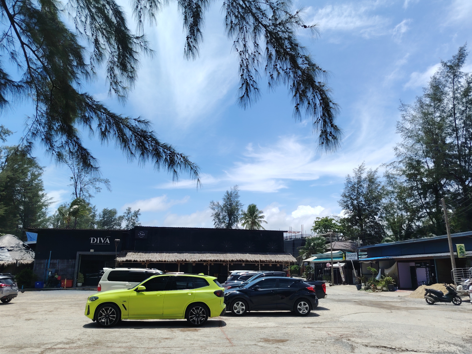 The image depicts a parking lot in front of a black building with the word "DIVA" in white lettering, located on Bang Tao Beach in Choeng Thale, Thailand. A bright yellow SUV is parked in the foreground, with a black SUV behind it. A white van is also visible in the parking lot. Trees and palm trees line the background, with a bright blue sky and scattered white clouds overhead. The building appears to be a restaurant or establishment with outdoor seating. A small motorcycle is parked to the right of the frame. The overall scene suggests a sunny day in a tropical coastal environment.