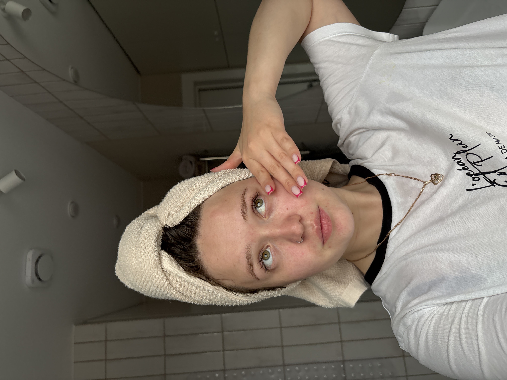 A young woman is in a bathroom, likely in a private residence. She has a towel wrapped around her head, suggesting she recently washed her hair or face. She is wearing a white t-shirt with black trim and is also wearing a gold necklace with a heart pendant. Her nails have pink tips. She is touching her face near her left eye with her right hand. The bathroom has gray tiled walls and floor. The image may be taken in Izhevsk, Russia, but there are no visual markers to confirm the location.