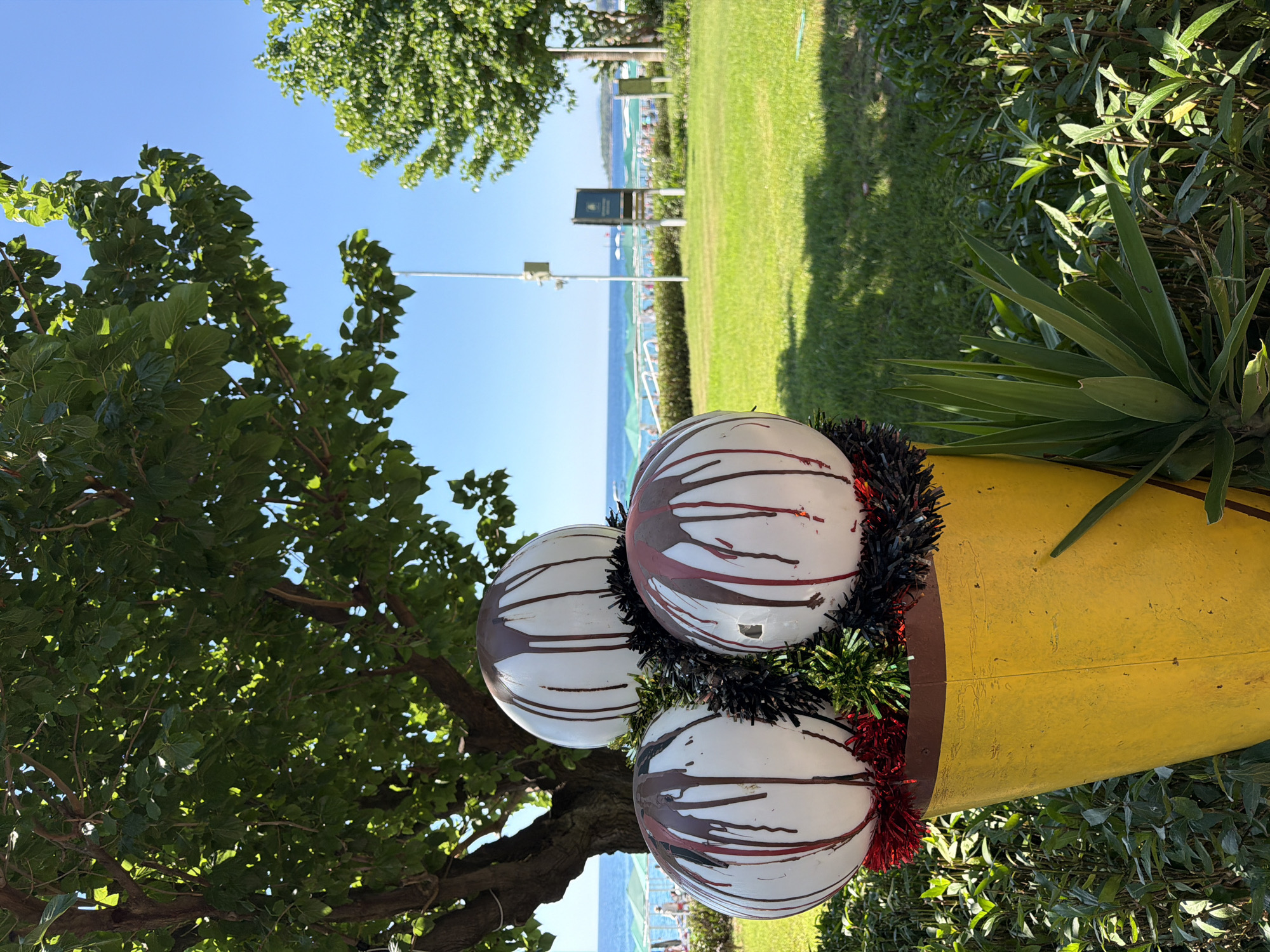 In an outdoor setting under a clear blue sky, a whimsical ice cream cone sculpture is the focal point. The sculpture, designed to mimic a cone, is a large yellow conical container filled with three white spherical "scoops" of ice cream. These scoops are adorned with drips of dark brown, resembling chocolate sauce, and some with streaks of red. The "ice cream" is further decorated with black and red tinsel.
Behind the sculpture, a lush green lawn stretches towards a body of water, likely the sea, visible in the distance. The shoreline is lined with numerous beach umbrellas, suggesting a public beach. Several people can be seen lounging or walking along the beach. Trees with dense green foliage frame the scene, casting dappled shadows on the grass. In the background, a white utility pole and a dark informational sign are present. The environment appears to be a well-maintained park or resort area situated by a beach.