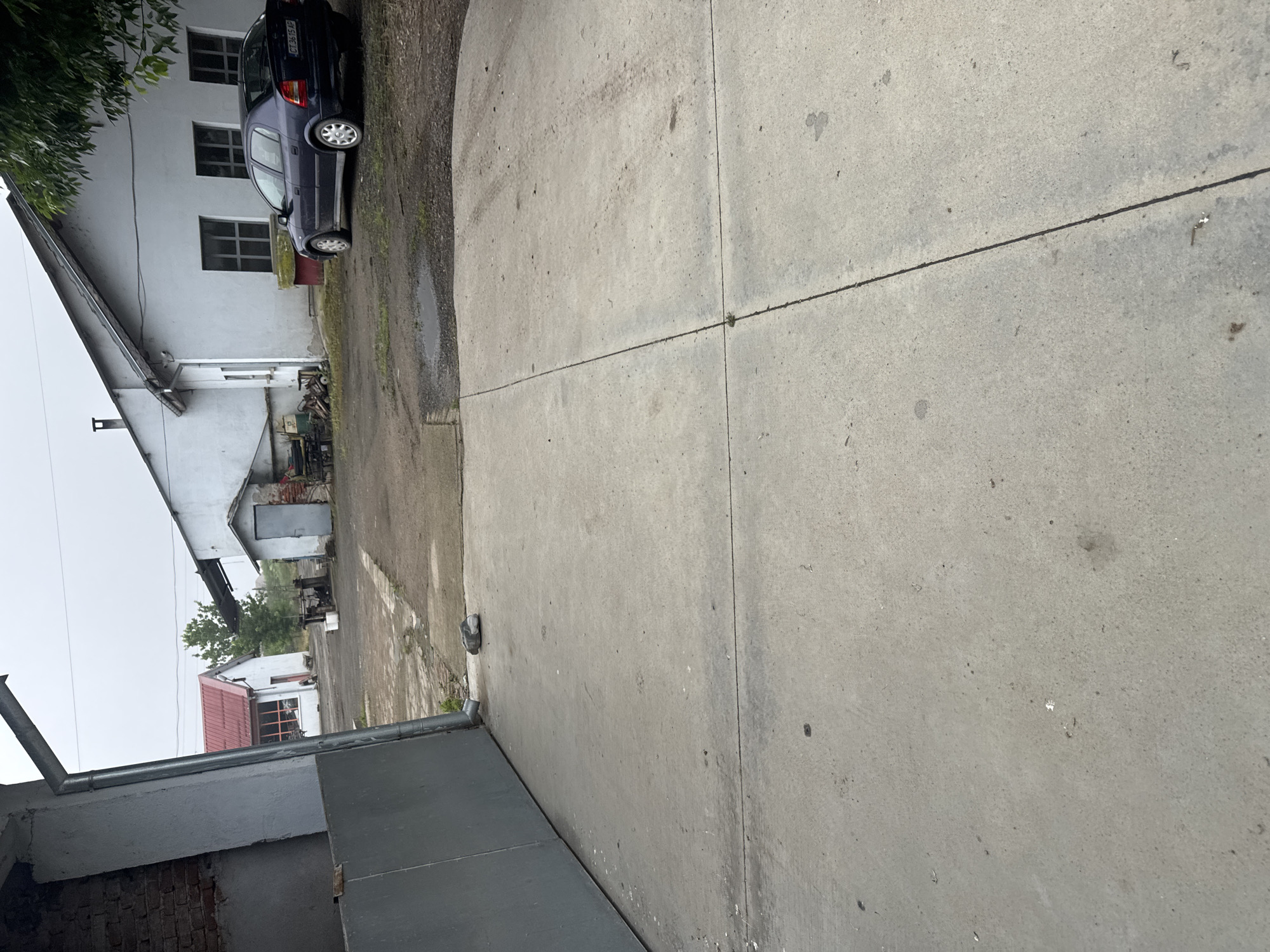 This image depicts an outdoor scene, likely a yard or driveway in Sarnevo, Bulgaria, under an overcast sky. The foreground and right side of the image are dominated by a light grey concrete path, divided by dark expansion joints. The concrete surface shows various stains, marks, and scattered specks.
To the left, a dark blue hatchback car, resembling an Opel Astra, is parked on a dirt and gravel area. Its rear license plate, "CC 0315 AP", is clearly visible. The car is situated next to a plain, two-story white building featuring several dark-framed windows. The upper part of this building's roof and some surrounding green tree foliage are visible on the far left. A chimney is also noticeable on the lower portion of the building towards the left.
Further into the background, additional white structures can be seen, including a building with a distinctive red-tiled roof and a large window or door, partially obscured by other objects and light green foliage. In the lower-left corner, modern metal guttering and a downspout are prominent, attached to an unfinished-looking building section that exposes some red brickwork.
The lighting is soft and diffused due to the overcast conditions, indicating daytime. Wet patches on the ground suggest recent rain or general dampness. No people or specific activities are visible, presenting a static, quiet rural or semi-rural setting.