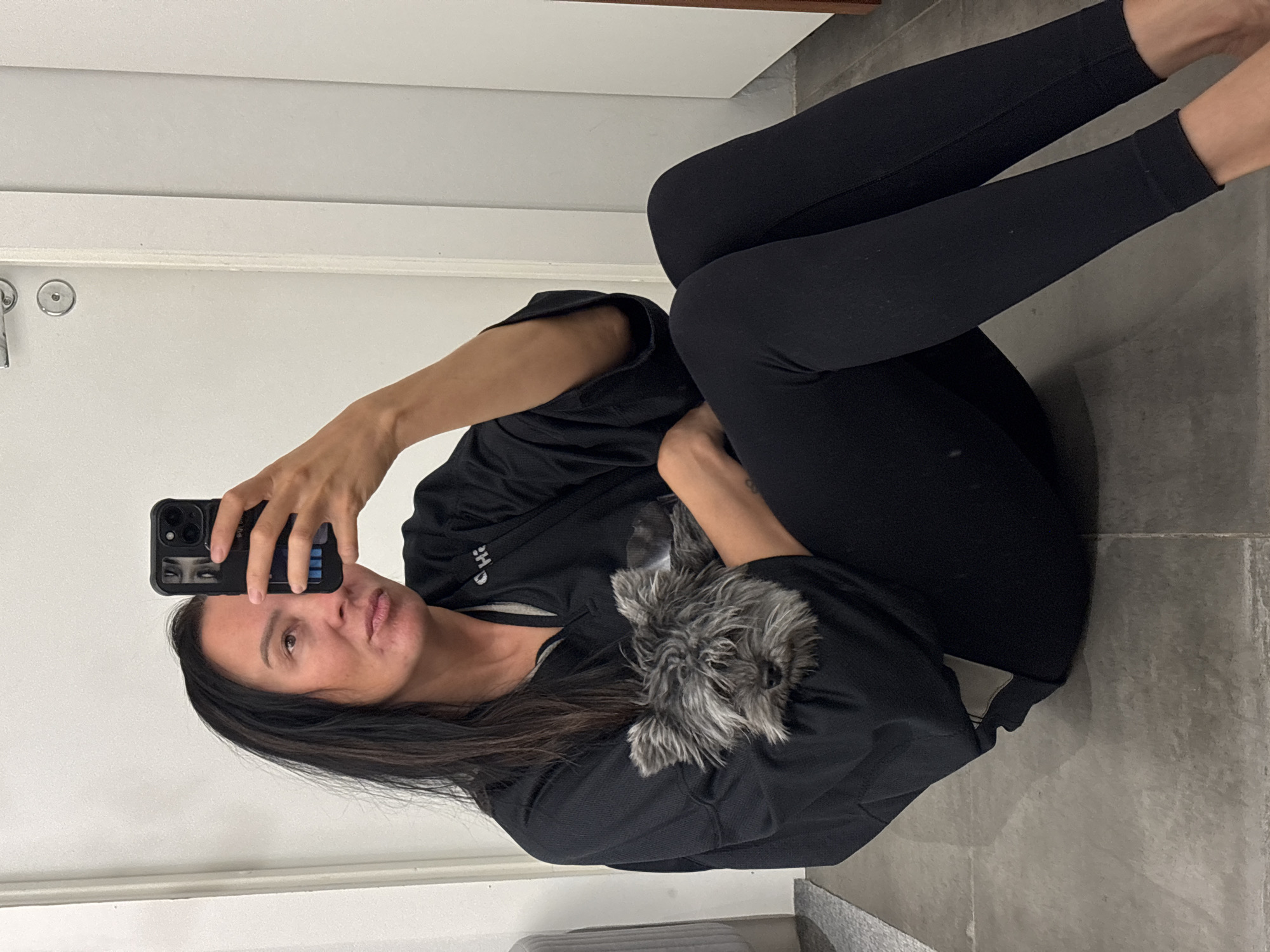 A woman wearing black leggings and a black shirt sits on a gray tiled floor, holding a phone in her right hand to take a selfie. A small, gray, fluffy dog sits in her lap. The background consists of a white wall with a door and silver handle. The location cannot be determined from the image.