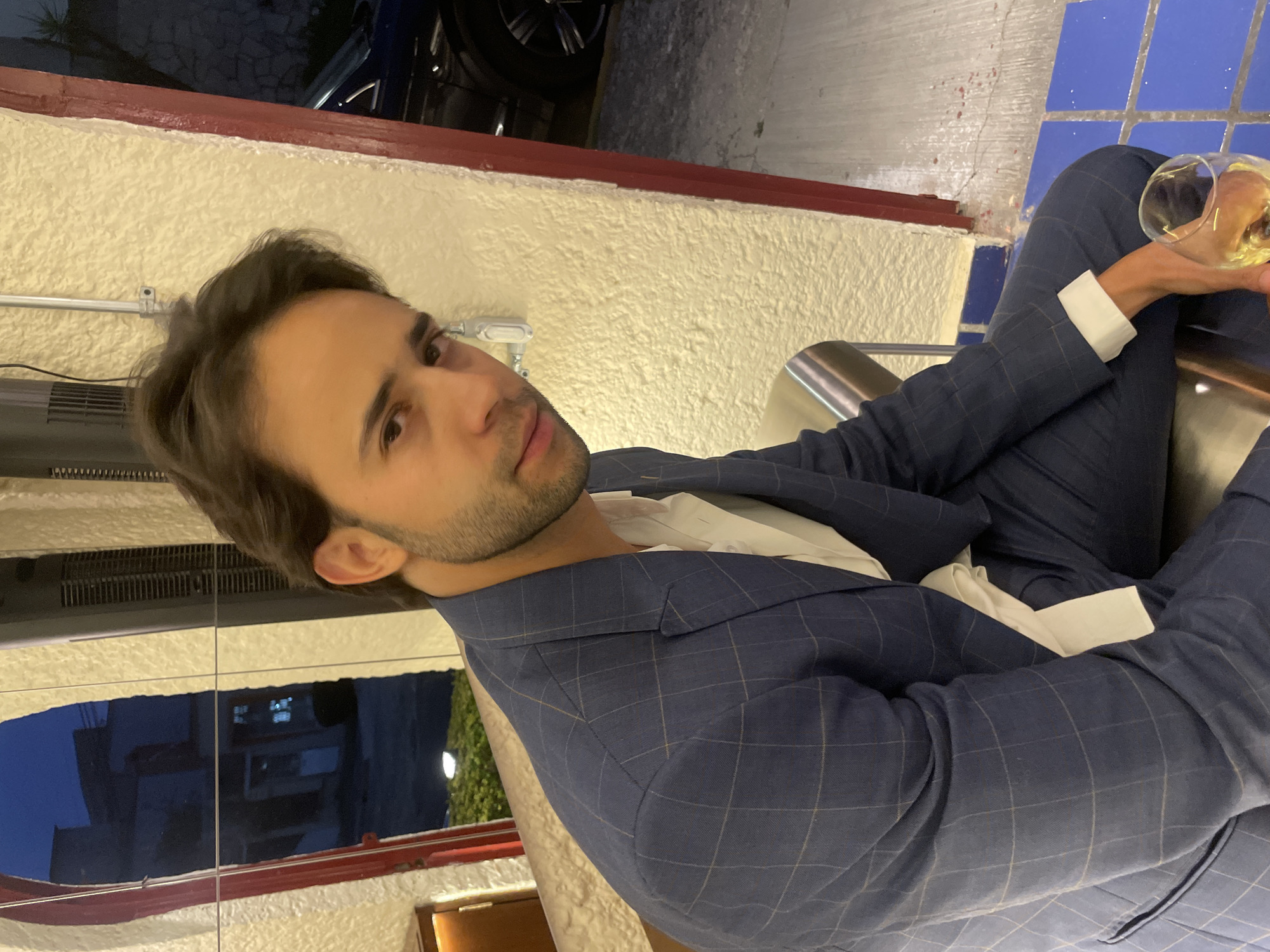 The image shows a man in a blue patterned suit, white shirt and white cuffs, seated and holding a glass of clear beverage, likely wine. He is looking at the camera. Behind him, there is an interior wall and a glimpse of an exterior scene, possibly a patio or outdoor dining area, with a dark blue structure visible in the upper left corner and light-colored masonry walls. The lighting suggests it is either evening or an artificially lit interior. The style of the building's architecture, particularly the tile work, is typical for Puebla, Mexico. The man is in a formal setting, possibly a restaurant or bar in Puebla.