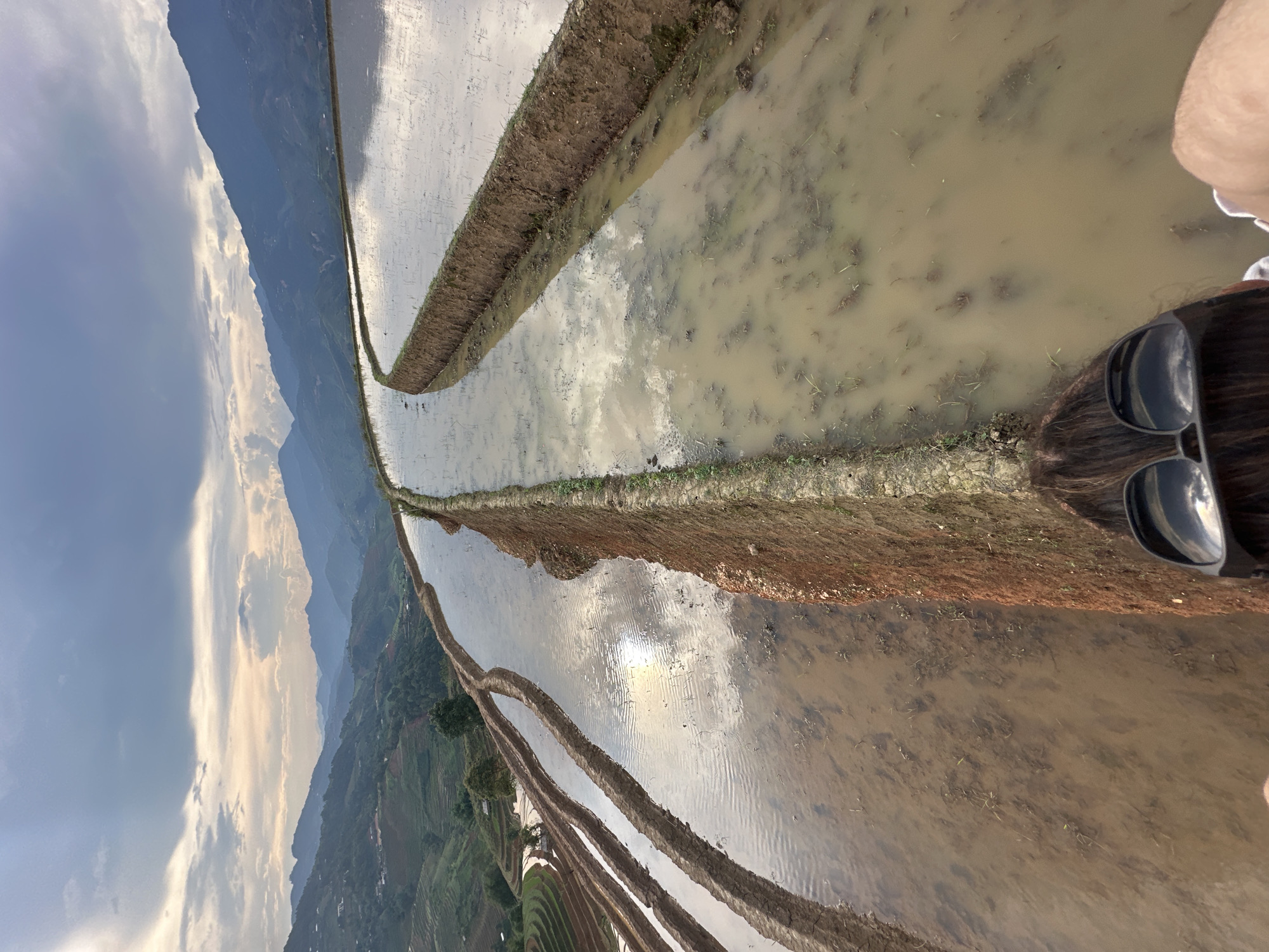 This image, captured in Lao Chai, Vietnam, presents a dramatic, vertically oriented view of extensive rice terraces nestled within a mountainous landscape. In the top right corner, partially visible, is the head and shoulder of a person with dark hair, wearing black sunglasses, suggesting they are looking down at the scene.
The central focus is on multiple tiers of water-filled rice paddies, separated by narrow earthen dikes. The surface of the water brightly reflects the sky above, showcasing intricate patterns of clouds and a luminous spot that appears to be the sun's reflection. The water in the paddies closest to the viewer is somewhat turbid, with visible submerged mud and sparse aquatic vegetation.
Beyond these foreground terraces, the landscape unfolds into a deep valley dotted with more terraced fields and lush green vegetation on the distant slopes. Majestic mountains rise in the background, their dark forms contrasting with the expansive sky. The sky itself is largely cloudy, but bright, suggesting a late afternoon or early morning setting with diffused light. The overall atmosphere is serene and highlights the impressive scale of human-shaped agricultural terrain within a grand natural setting. No visible text is present in the image.