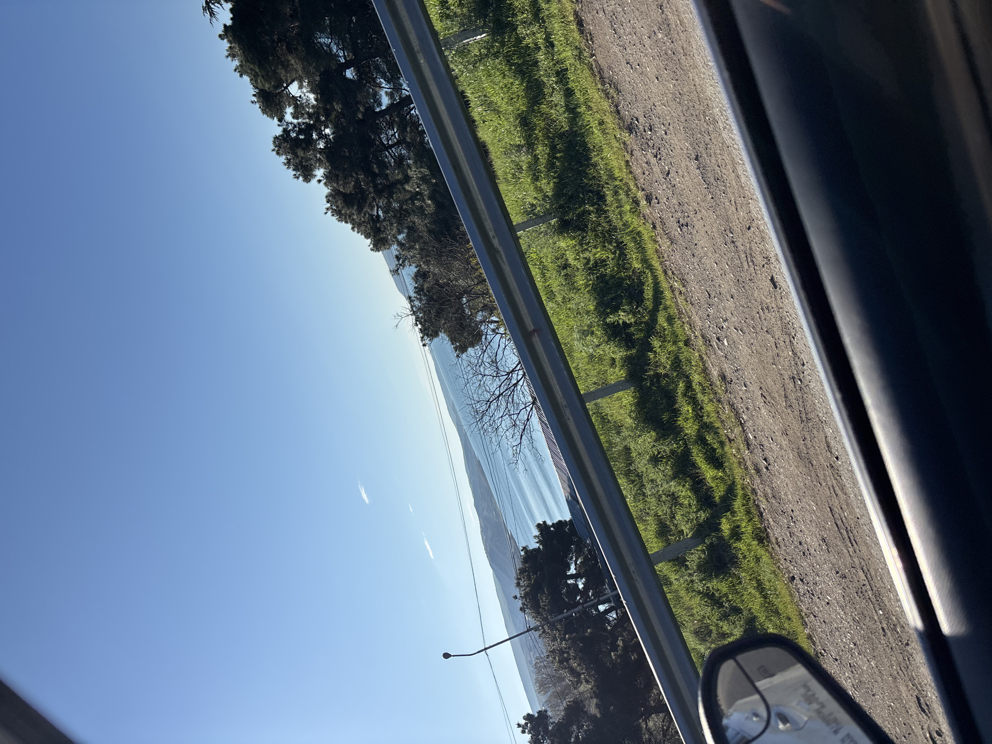 The image is taken from inside a moving vehicle. Outside the window, there's a clear blue sky overlooking a large body of water, possibly a lake or reservoir, bordered by a visible coastline. A guardrail is situated between the road and the grassy area. Some trees are visible along the shoreline, along with an electrical pole. The rearview mirror of the vehicle is partially visible. Based on the landscape and context clues, the location is likely near Tbilisi, Georgia, potentially close to the Tbilisi Sea or other reservoir in the area. The photo captures a roadside scenic view.