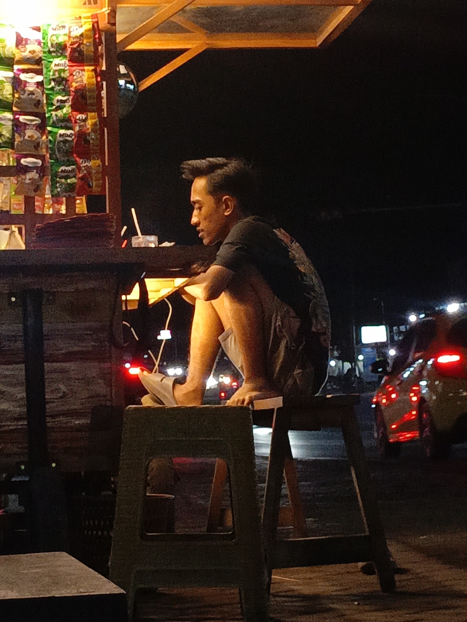 A young man is sitting on a wooden stool at night in Purwoharjo, Indonesia. He is wearing a dark t-shirt and light-colored shorts, with grey flip-flops. His legs are bent and he is holding something in his hands, possibly an item from the stall he is near. The stall, visible to the left, is stocked with numerous packaged goods, appearing to be snacks or drinks. In the background, out-of-focus car lights suggest a street scene with traffic. The lighting is dim, with warm light illuminating the stall and the man.