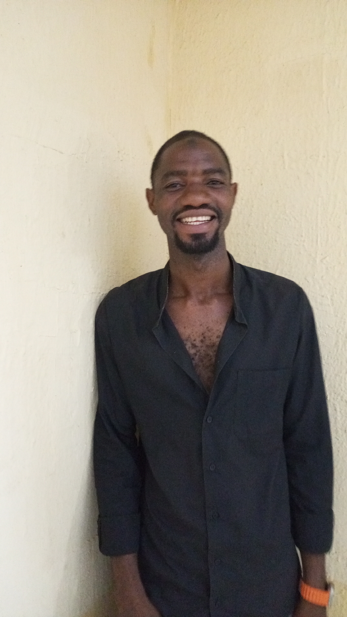 A man stands indoors against a pale yellow wall, possibly stucco. He is Black, smiling, with a beard. He wears a black long-sleeved shirt, partially unbuttoned. He has an orange wristband on his left wrist. The location is likely Sabon Tasha, Nigeria.