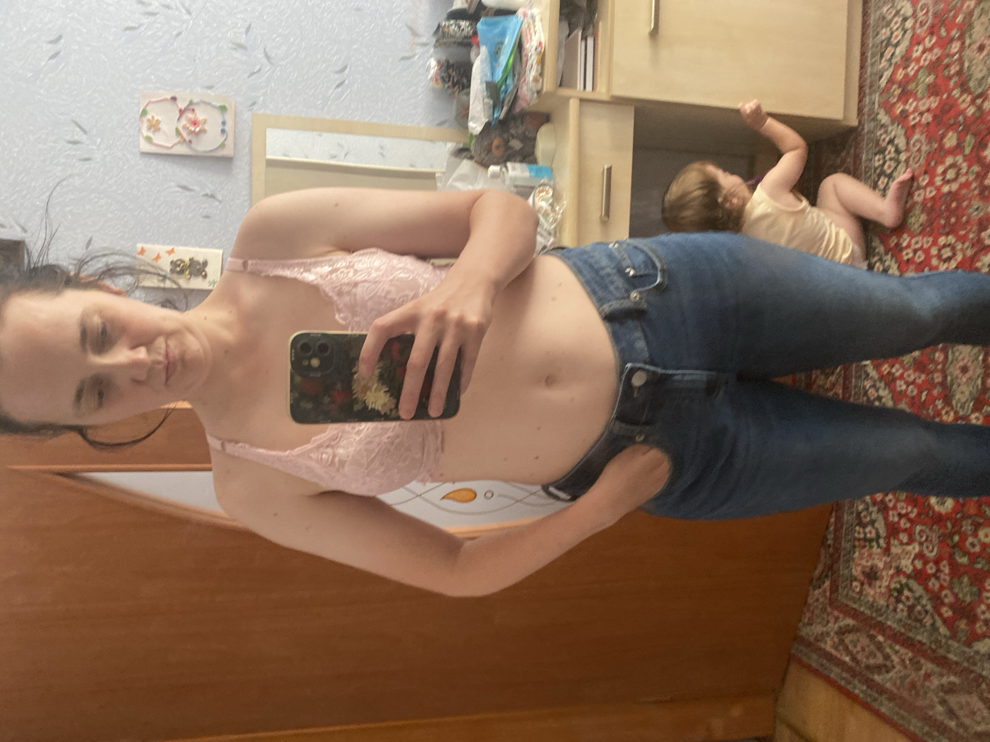 In an indoor setting, a woman stands in front of a wooden door and a vanity mirror. She is wearing a pink lace bralette and blue jeans. She is holding a smartphone with a floral case in her right hand, and her left hand is in her pocket. A toddler is visible in the background, playing on a red patterned rug near the vanity. The room appears to be a bedroom or dressing area, with decorations on the wall behind the mirror.
