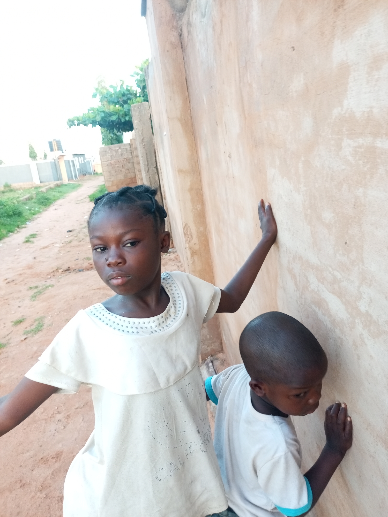 Two children are near a tan-colored wall. The child in the foreground is a girl wearing a white dress. She is looking at the camera with a serious expression. Her hair is braided into cornrows. The child in the background is a boy wearing a white t-shirt with blue trim on the sleeves. He is leaning against the wall and looking down. The background shows a dirt path, some green vegetation, and a building. The location is unknown.