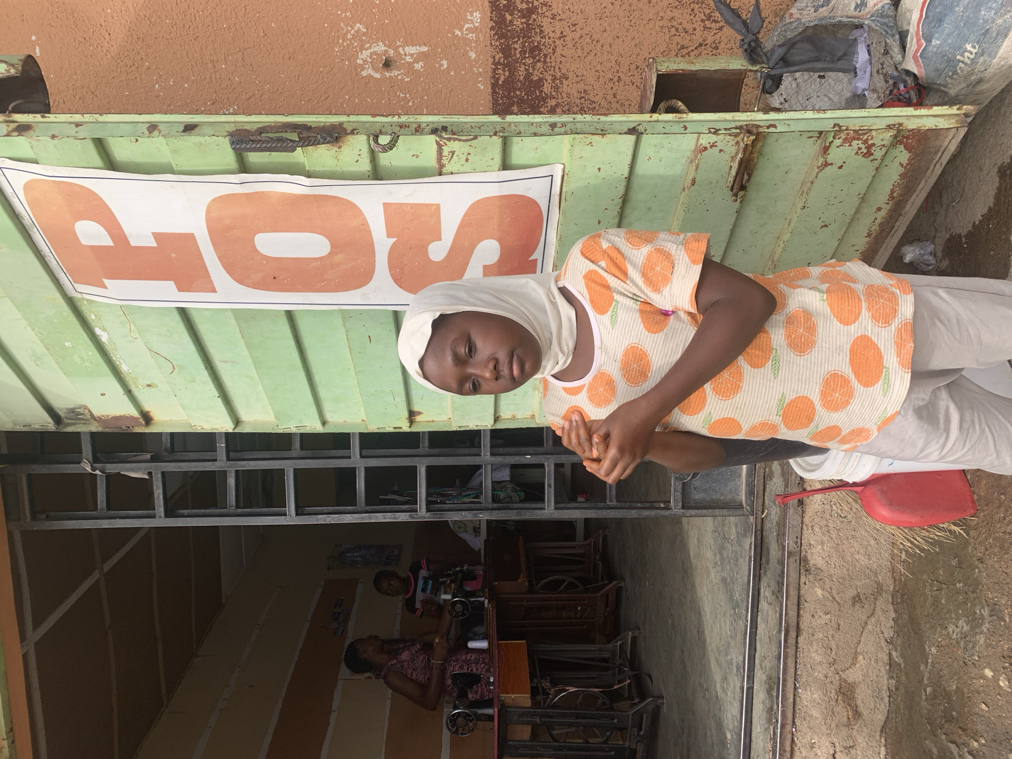 An outdoor scene in Jos, Nigeria, features a young girl standing in front of a weathered, green corrugated metal structure, likely a kiosk or workshop. The image is oriented vertically, showing the girl centered with the structure behind her.
The girl wears a light-colored head covering and a short-sleeved top patterned with orange citrus slices and green leaves. Her hands are clasped together in front of her. Her expression is calm and neutral as she looks towards the viewer.
Affixed to the top part of the green structure is a white banner with large orange letters, clearly displaying the word "STOP". Above the green structure, a textured, reddish-brown wall is visible. To the girl's right, a red dustpan and a broom rest on the ground.
Behind the girl, through an open doorway or window of the green structure, an interior workspace is visible. Inside, two adult women are seated at what appear to be traditional, foot-powered sewing machines, suggesting a tailoring or textile workshop. The interior features metal framing and simple wooden tables.
The ground around the structure is rough concrete or dirt. The overall setting appears to be a bustling, everyday street or market environment, with natural light indicating daytime. The structures show signs of age and wear.
