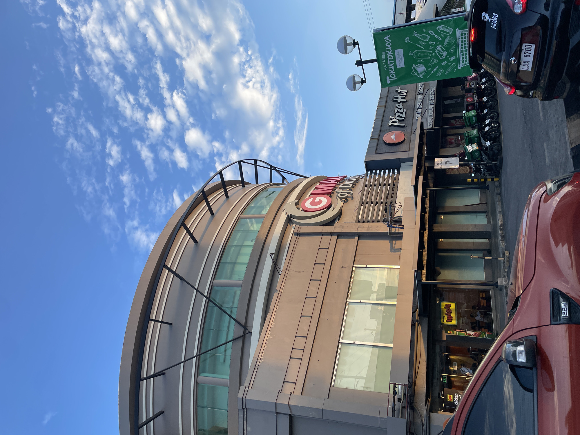 This image shows a street view in Davao City, Philippines, under a bright blue sky with scattered white clouds. The scene features a modern, multi-story building with a curved facade, primarily clad in grey and glass. A prominent red and white circular logo with the letters "GMA" is visible on the building's exterior.
Adjacent to this building is another establishment with a sign that reads "Pizza Hut" and a green banner displaying various kitchen utensils and the text "Tokutokuya From Japan." A line of parked motorcycles, many with green "Grab" branding, suggests a delivery hub or a busy commercial area.
Several vehicles are parked in front of the establishments. A black Nissan sedan with a license plate "LAA 8700" is parked facing away from the camera, and the rear of an orange vehicle is visible in the foreground, partially obscuring the street. Text visible includes "EXIT," "Chicken," and "NO PARKING." The lighting indicates it is daytime, possibly late afternoon, given the angle of the shadows. The overall atmosphere is that of a commercial district with active food services and delivery operations.