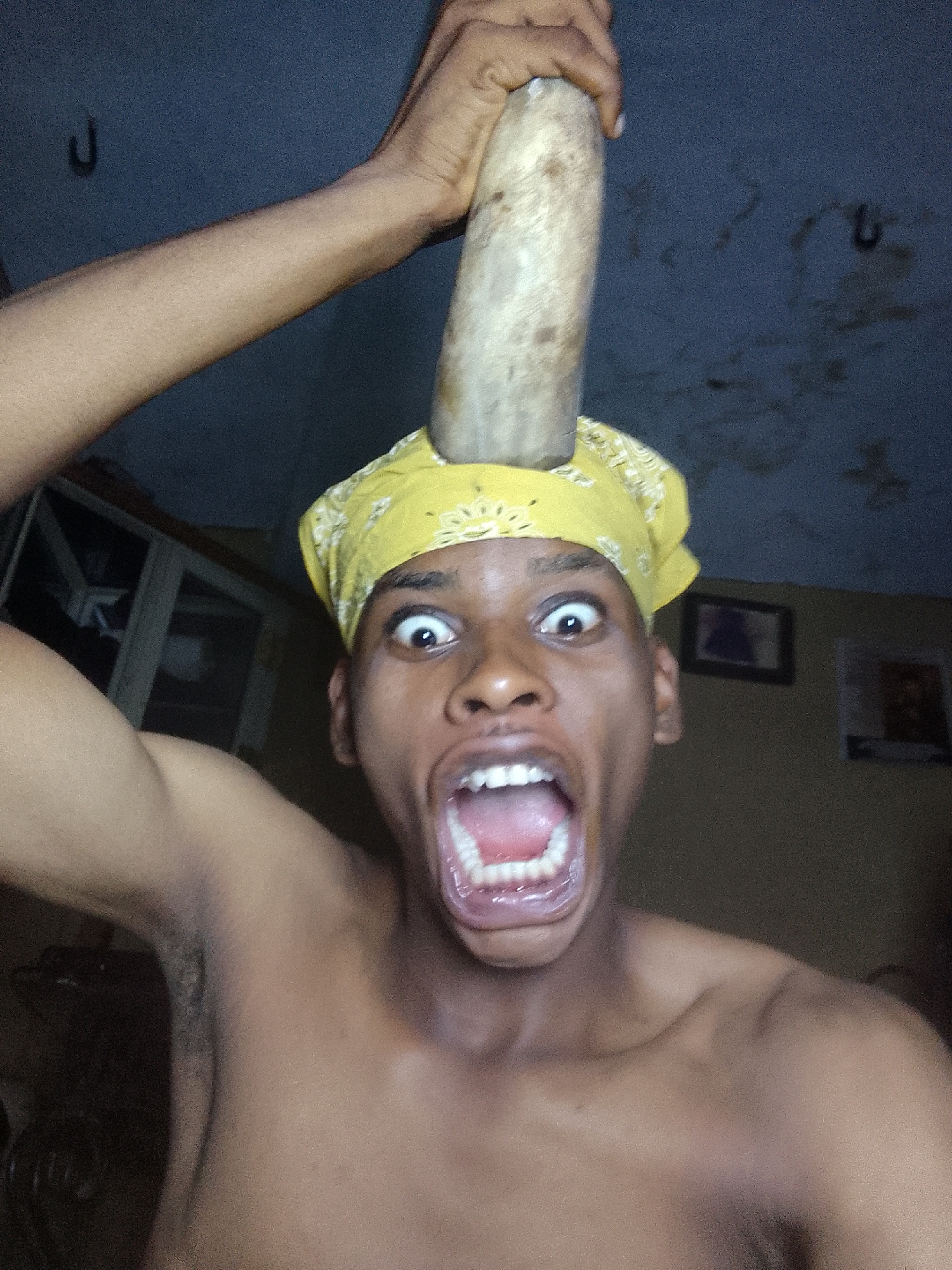 A shirtless, dark-skinned man with a yellow bandana is in the foreground. He is looking at the camera with wide eyes and an open mouth, conveying a look of shock or surprise. Atop his head, balanced on the bandana, is a thick, cylindrical object with a light, possibly wooden appearance. The man's right arm is raised above his head, holding the object in place. His left arm is extended to the side.
In the background, the wall is a light beige color and features a framed picture. A dark object resembling a hook is attached to the ceiling. In the corner of the room, there appears to be a glass cabinet or display case with partially visible contents.
The location is in the city of Unknown, Unknown.