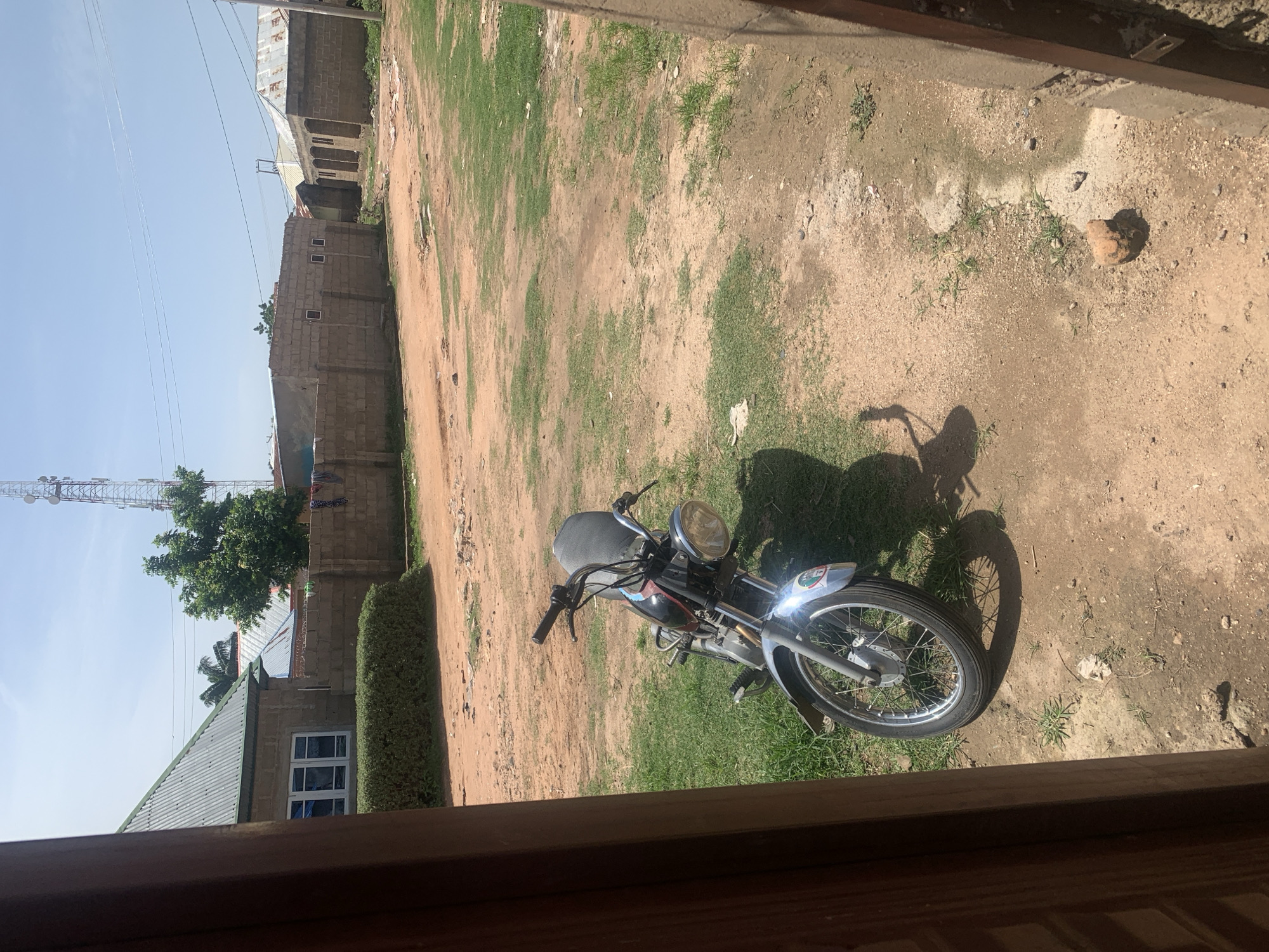 In Jauro Yinu, Nigeria, a motorcycle is parked on a patch of grass next to a dirt and gravel area. The motorcycle has a black seat, a silver front fender, and chrome handlebars. Behind the motorcycle, a low, green hedge runs along the base of a brick wall. Beyond the wall, there are several buildings constructed of concrete blocks with corrugated metal roofs. One building on the left has a green metal roof and a white-framed window. Further in the background, a tall communications tower with multiple platforms stands against a clear blue sky, with power lines visible between the buildings and the tower. The overall scene depicts a rural or semi-urban environment under a bright sun.