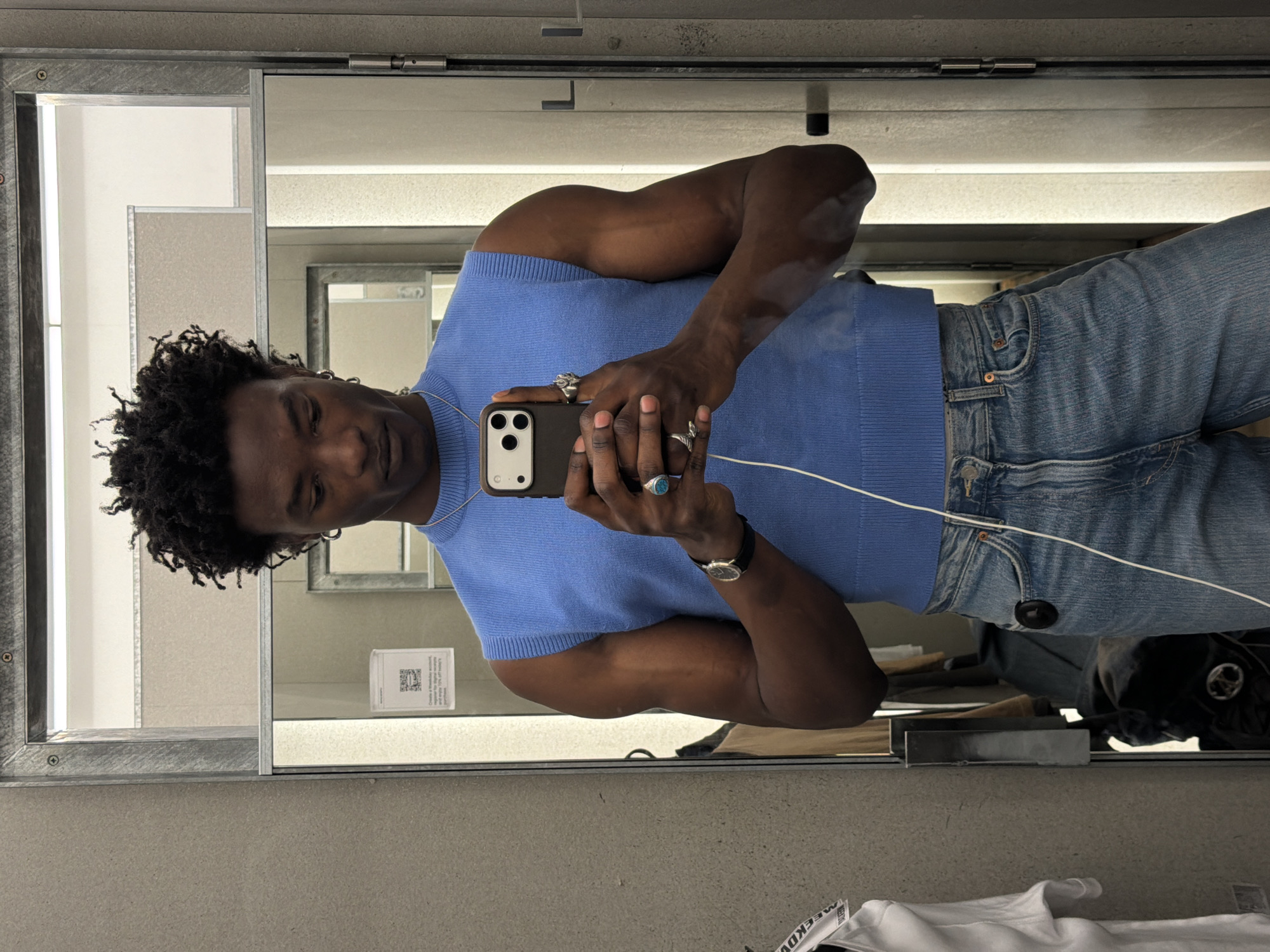 A dark-skinned man with short, curly black hair takes a mirror selfie, holding a dark-colored smartphone with a white cable connected to it. He is wearing a light blue sleeveless top and blue denim jeans. His eyes are focused downwards, presumably on the phone screen.
He accessorizes with several rings: a large silver-toned ring on his right index finger, another silver ring on his right ring finger, a turquoise-colored stone ring on his left index finger, and a silver ring on his left middle finger. A thin silver chain is visible around his neck, and a watch with a dark strap and a light-colored face is on his left wrist.
The selfie is taken in what appears to be a changing room or retail environment, suggested by the large mirror with a metallic frame. The reflection shows light-colored walls in the background. In the actual foreground, a grey wall is visible, and on the right, a white garment with a black tag bearing the text "MEEKDV" is present. A black security tag is also visible hanging from his jeans in the reflection. On the mirror's surface, a small white sign with a QR code and text, partially readable as "Create a free account...", "exclusive offers", and "enjoy 10% off", is reflected. The setting is a casual retail space in London, United Kingdom.