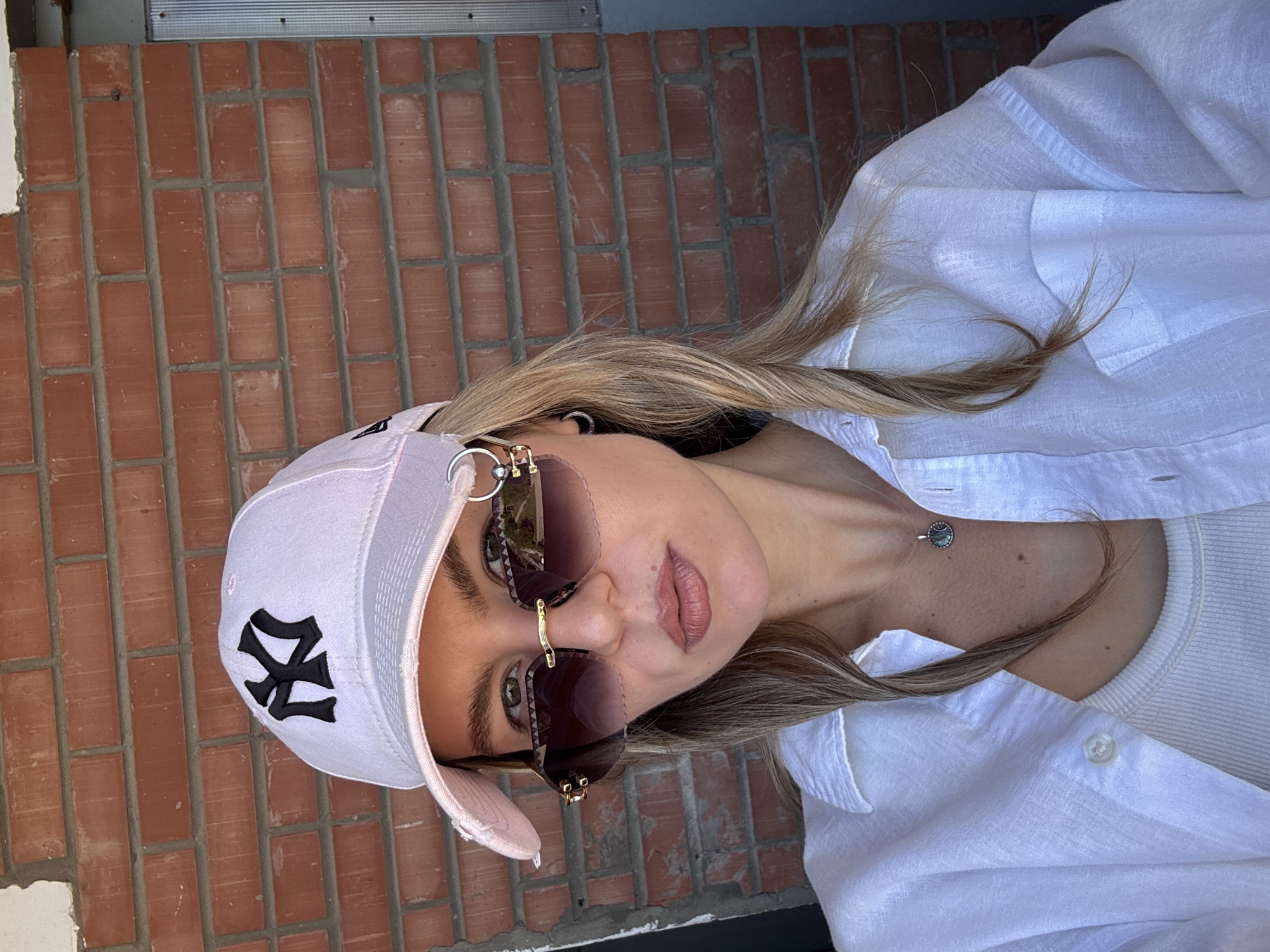 The image shows a close-up of a person wearing a light pink baseball cap with a black logo, large sunglasses with dark lenses and gold accents, and a white button-down shirt over a white ribbed top. The person has blonde hair and is wearing subtle makeup. A thin necklace with a circular pendant is visible on their neck. The background is a red brick wall. The lighting suggests it might be daytime, possibly outdoors. The image is taken from a slightly high angle, looking down at the subject. The overall impression is casual and stylish. The logo on the cap appears to be the "NY" logo, commonly associated with the New York Yankees baseball team.