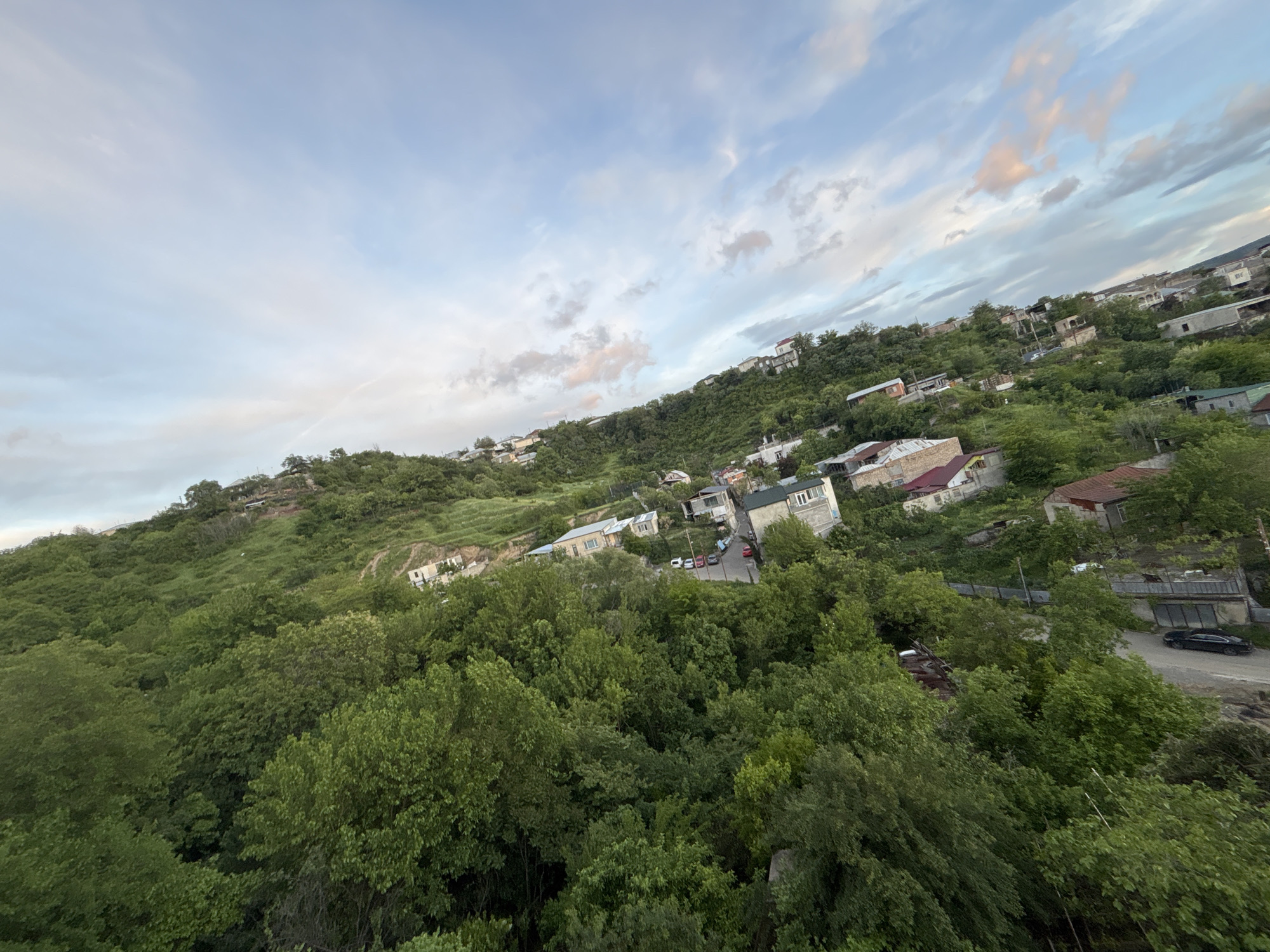 The image depicts a hillside residential area, likely located on the outskirts of the city of Tbilisi, Georgia. The scene is dominated by a steep slope covered in dense green trees and vegetation, with houses and buildings interspersed amongst the trees. The housing styles appear varied, ranging from small, simple structures to more substantial buildings. A winding road is visible along the hillside, with a few cars parked. The sky is cloudy, with shades of white and blue dominating the upper portion of the image. The overall perspective suggests the image was taken from an elevated position, possibly a higher point on the hillside or from a building.