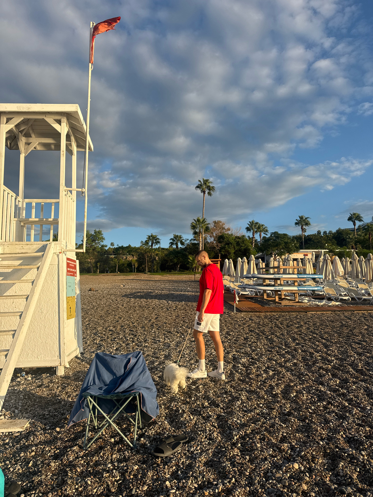 This image depicts a man walking a small white dog on a pebble beach in Antalya, Turkey, near a lifeguard station. The lifeguard station, painted white, is on the left side of the frame, with stairs leading up to it. A red flag flies from a pole next to the station. The man is wearing a red t-shirt and white shorts, and the dog is on a leash. In the background, rows of lounge chairs and umbrellas suggest a resort or public beach area. Palm trees are visible in the distance, characteristic of the Antalya region. The sky is partly cloudy with blue sky peeking through. A blue folding chair, covered with a blue tarp, and a pair of black sandals are in the foreground near the lifeguard station.