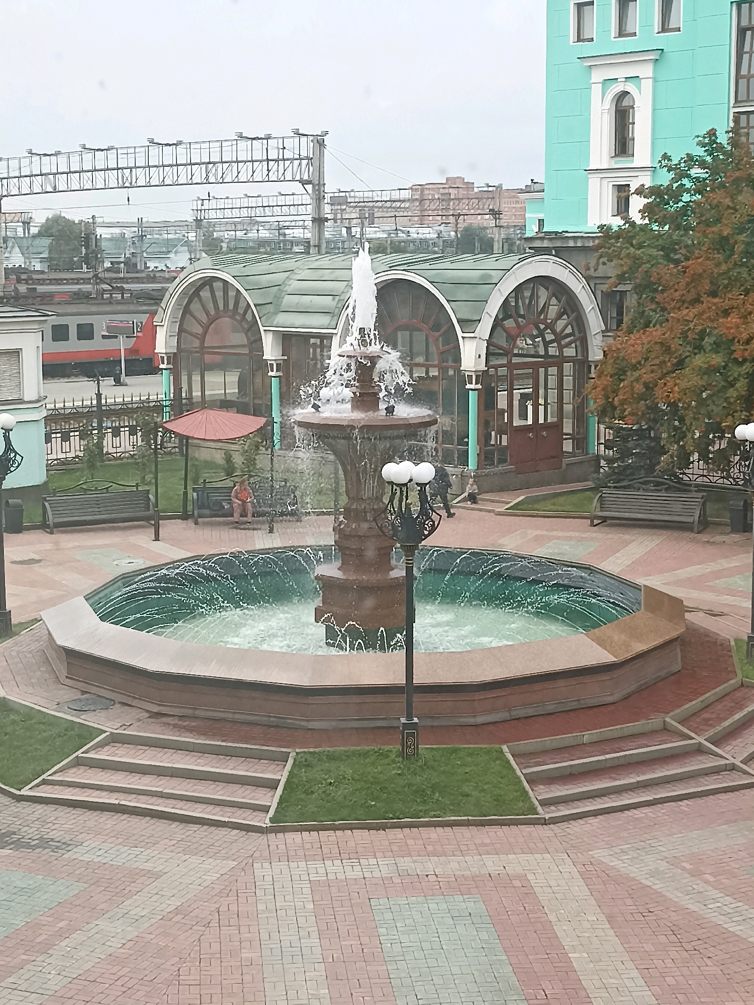 The image depicts a public square in front of a train station, identified as Novosibirsk-Glavny railway station in Novosibirsk, Russia. The central feature is a large, multi-tiered fountain with water spraying upwards. The square is paved with red and gray brickwork in a diagonal pattern. Steps lead up from the paved area to a grassy section surrounding the fountain.
In the background, a train with a red and white livery is visible on the tracks. Behind the fountain and to the right, a multi-story building with a turquoise facade and white trim stands. To the left of the turquoise building, a curved, arched structure with a green roof and large windows can be seen, part of the train station's architecture. Further in the distance, various industrial and urban structures are visible under an overcast sky.
Several benches are placed in the grassy areas. On one bench, a person wearing a light-colored garment is seated, holding a red object. Another bench to the left appears empty. A lamppost with multiple globes stands near the fountain, and another similar lamppost is visible further to the right. The overall scene suggests a moderately busy urban environment on a cloudy day.