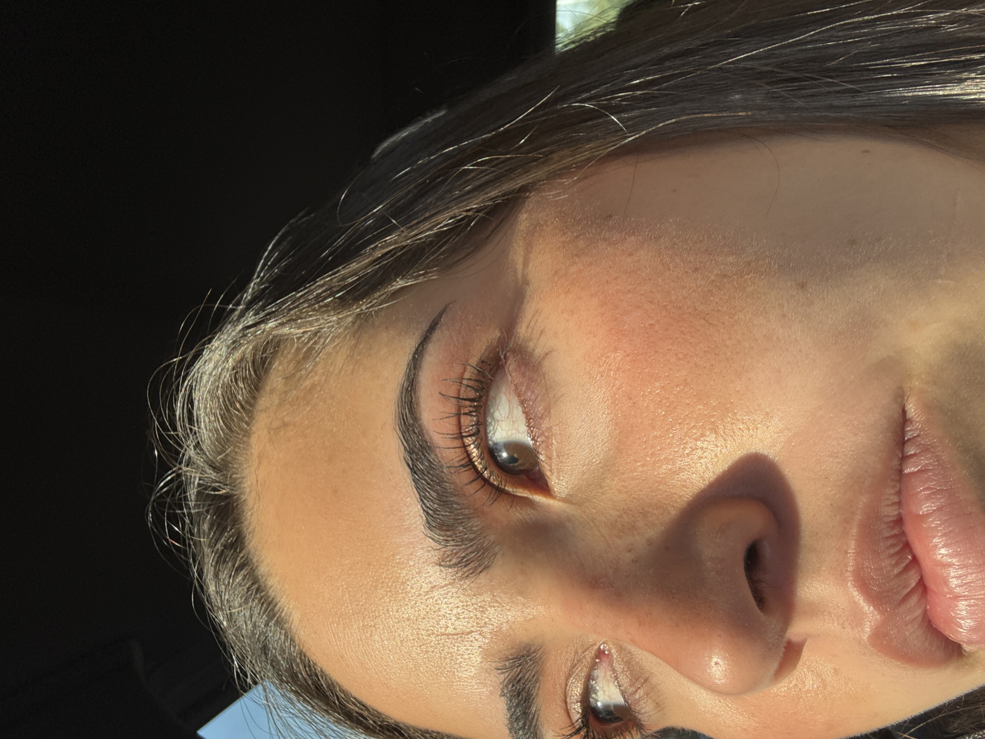 A close-up, sharply angled photograph shows a woman's face. The upper portion of her face is brightly illuminated, while the lower portion is partly in shadow. Her right eye is focused and visible, displaying long eyelashes and a defined eyebrow. The skin tone is fair, with slight visible pores. The lips are lightly colored, with visible texture. Her nose casts a clear shadow onto the adjacent cheek. The background is partially dark, with hints of light indicating an outdoor setting. It is impossible to determine the location in San Bernardino Tlaxcalancingo, Mexico without any distinct landmarks or contextual clues.