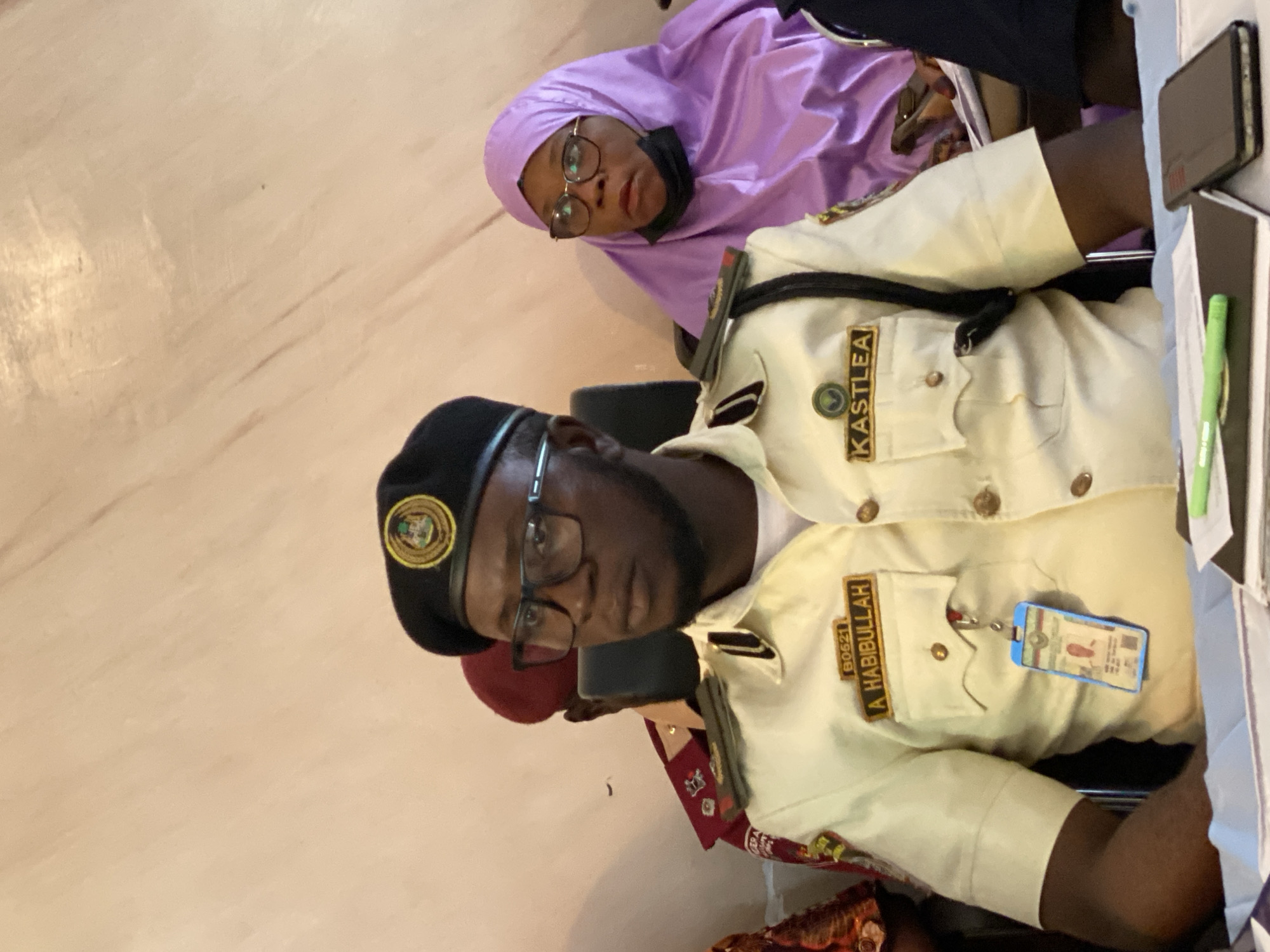 In a medium shot, a man wearing a tan uniform, black beret, and glasses is seated at a table. He has a name tag that reads "A. HABIBULLAH" on his left chest. A badge with a picture is hanging from his uniform. To his right, a woman wearing a purple hijab and glasses is also seated. A man in a red uniform is visible behind the man in the tan uniform. The background appears to be a light-colored wall. The scene is likely an indoor setting, possibly an office or meeting room in Unguwar Sarki, Nigeria, given the context provided.