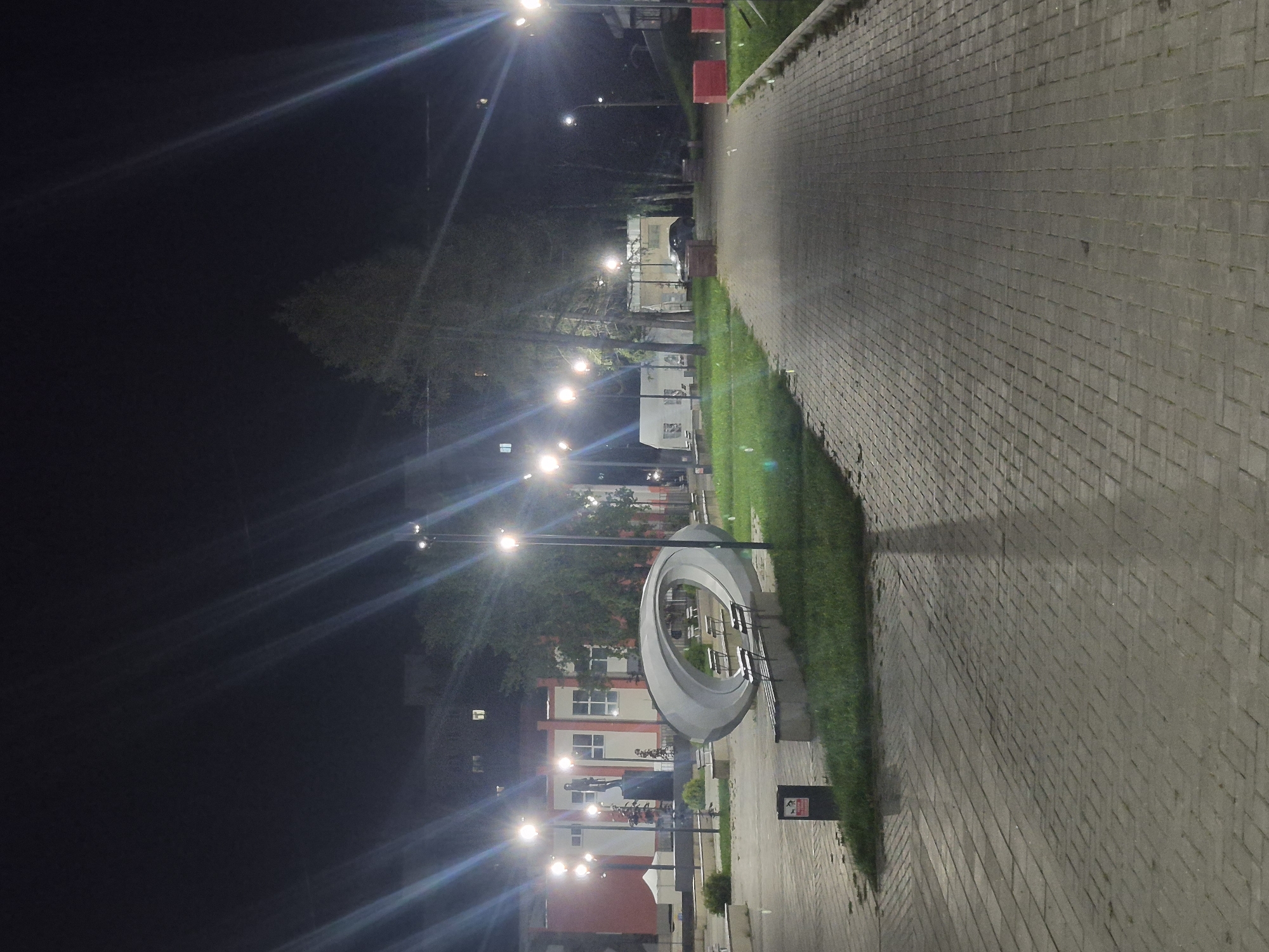 A paved walkway leads through a park at night. Streetlights illuminate the scene, casting long shadows. A modern sculpture, a gray, elliptical form, sits on a patch of grass to the left, with benches nearby. In the background, a building with a red facade and white windows is visible. A statue stands on a pedestal in front of the building. Trees and other foliage are scattered throughout the park. A dark-colored car is parked on the right side of the walkway. The ground is paved with gray bricks, and grass borders the walkway. Two red planters are visible on the right side of the walkway. The overall impression is a public space designed for recreation and art. This park is located in the city of Chaikovsky, Russia.
