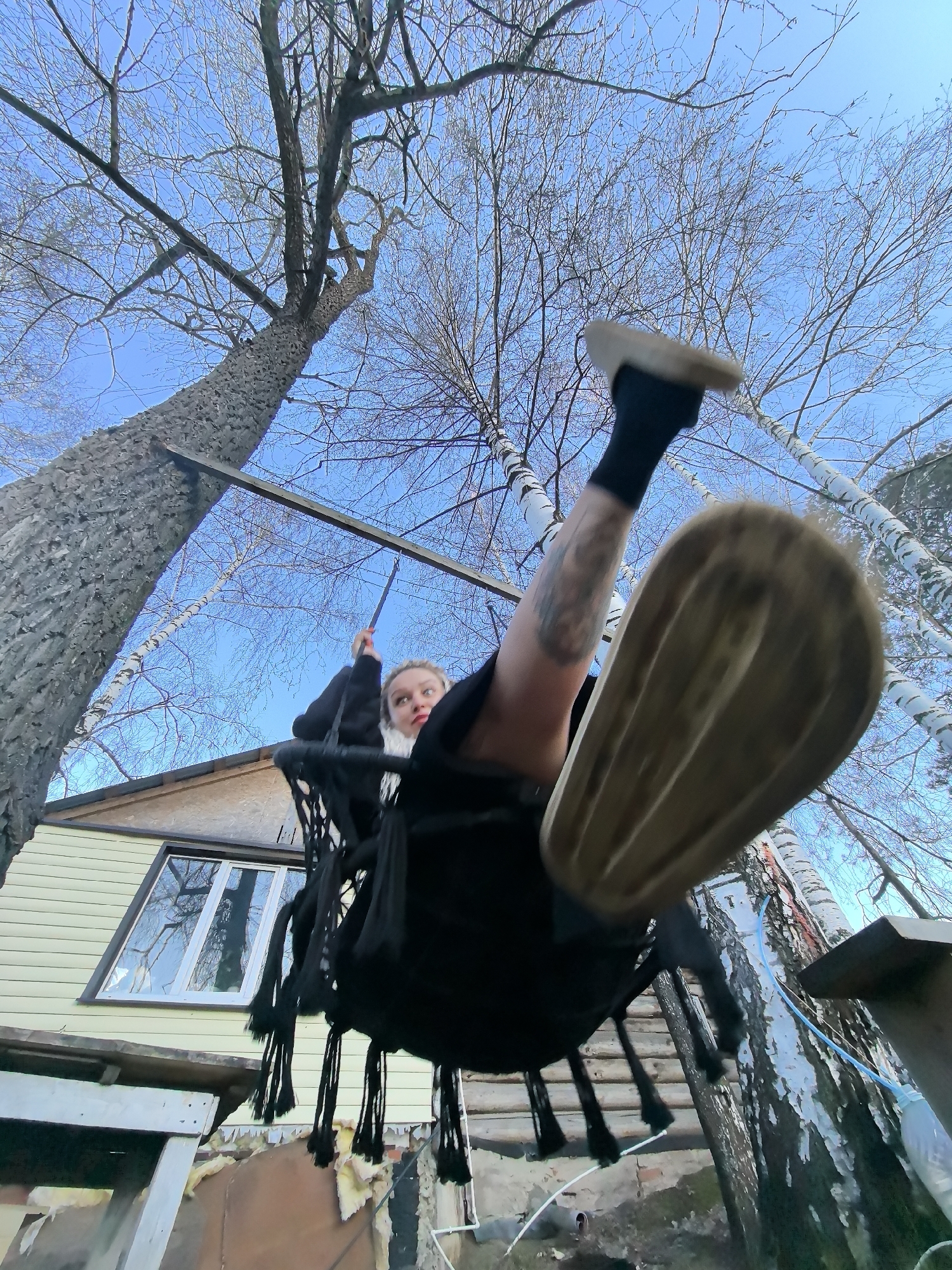 The image presents a dynamic, low-angle view of a person swinging outdoors in Noviy, Russia. A young individual, likely female, with light-colored hair, is shown in motion on a black, woven swing with decorative tassels. She is wearing dark clothing, including a black top or hoodie, dark shorts or a skirt, and black socks. Her light brown, platform-style shoes are prominently featured, with one foot extended towards the camera, creating a wide-angle effect and emphasizing the swing's momentum. Tattoos are visible on her left leg.
The swing is suspended from a metal or wooden beam secured to a large tree with thick, grey, textured bark, which anchors the left side of the frame. The background is dominated by numerous bare deciduous trees, primarily birches with their distinctive white bark, set against a clear, bright blue sky. This lack of foliage suggests the season is either late autumn or early spring.
In the mid-ground, a light yellow-beige house with a large, white-framed window is visible. The window reflects the sky and the surrounding trees. Below the house, some debris and exposed insulation material can be seen. A light blue hose or tube runs vertically near one of the birch trees. The scene captures a moment of playful activity in a natural, somewhat rustic residential setting. There is no visible text in the image.