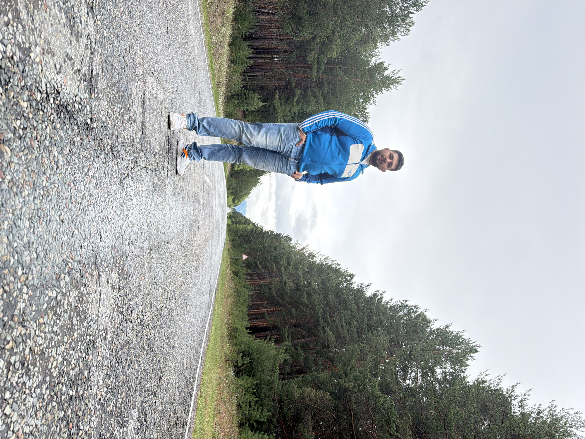 A man stands on a paved road. He is wearing jeans, white sneakers with orange accents, and a blue jacket with white stripes. The road is bordered by trees. The location is Сизая, Russia. The sky is overcast.