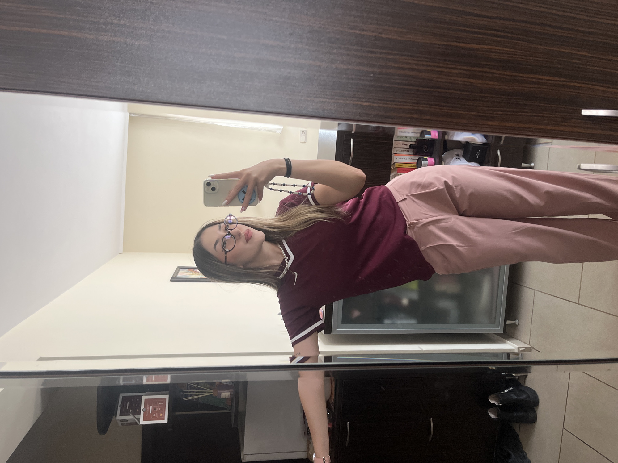 The image, taken in Sofia, Bulgaria, shows a young woman capturing a mirror selfie indoors. She is positioned facing a large mirror, holding a silver iPhone with a light-colored case in her right hand to take the photo. The woman has long, light brown hair and is wearing round-framed glasses. She is dressed in a maroon short-sleeved top with white striped cuffs and a small white Nike logo on the chest, paired with light pink, wide-leg trousers. A black bracelet is on her left wrist, and a light-colored smartwatch is on her right, while she also wears a pearl-like necklace. Her fingernails are painted white.
The reflection in the mirror reveals the room's interior. On the left side of the reflection, a cream-colored wall features a small framed picture, with various items on a dark surface below it, including a prominent red-brown box. On the right, a dark brown wooden shelving unit or wardrobe is visible, containing stacked books and other objects. Some of the book titles, though mirrored and difficult to read, appear to include "HYLE", "ZMAD", and "SEZAM". A white electrical outlet is also seen on the wall. The floor is covered with light-toned rectangular tiles. In the foreground, below the woman's reflection, a dark cabinet is visible, with a pair of dark shoes resting on the floor in front of it. The scene suggests a casual moment within a personal, well-appointed living space.