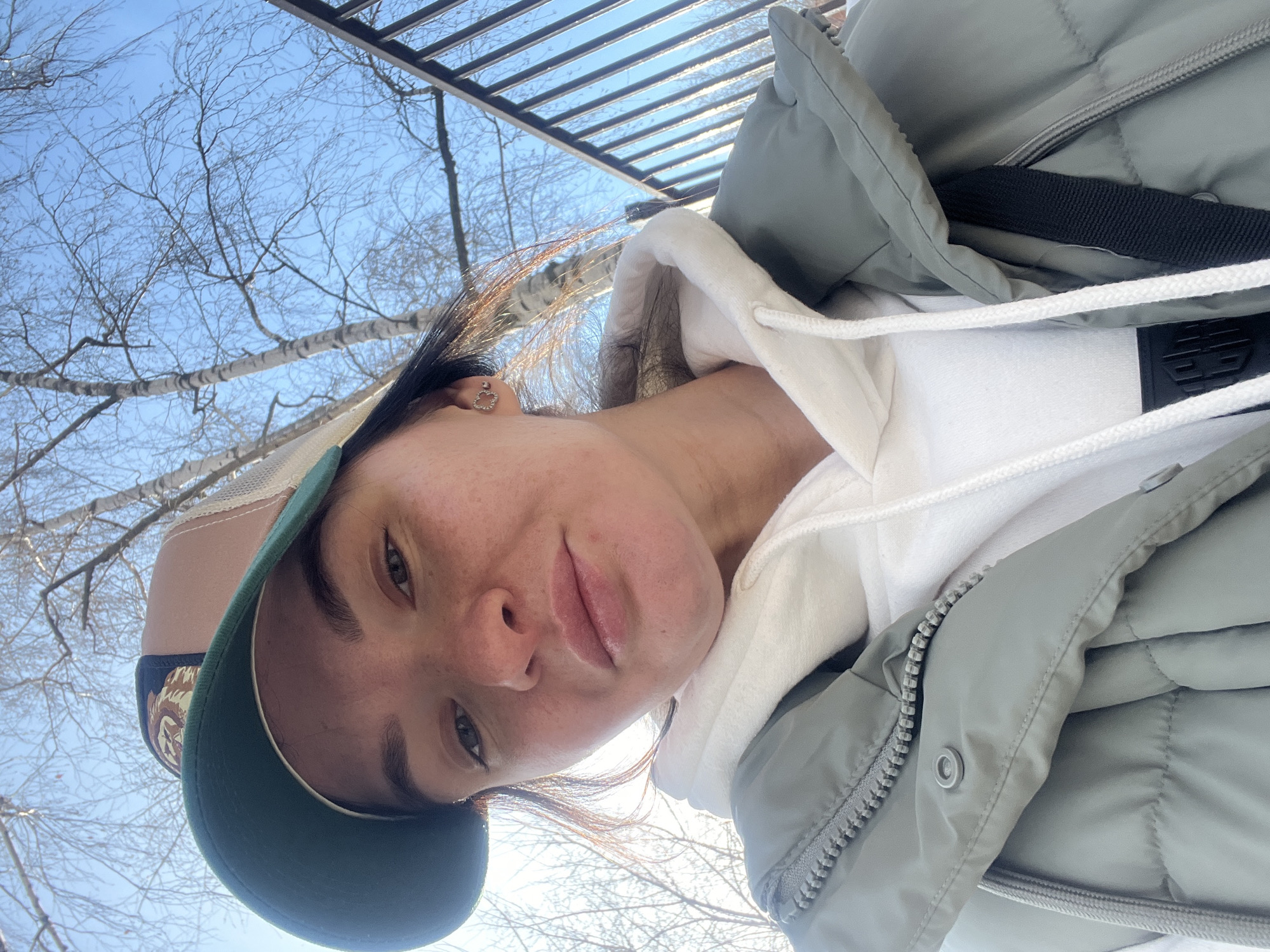 A woman wearing a green and tan baseball cap, a white hoodie, and a light gray puffer jacket is positioned in the lower-left quadrant of the frame. She is looking directly at the camera with a neutral expression. Visible freckles are scattered across her nose and cheeks. Bare tree branches and a segment of black metal fencing are visible in the background, against a pale blue sky. The location cannot be definitively determined as Kazan, Russia, from the provided image.