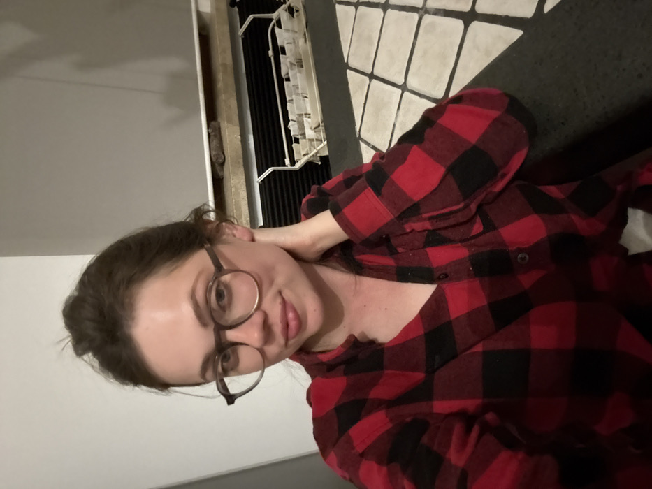 The image displays a single female subject in an indoor setting. The woman possesses light skin, dark hair pulled back into a bun, and wears round eyeglasses with dark frames. She is dressed in a red and black plaid long-sleeved shirt, partially unbuttoned at the collar. Her right hand is positioned at the back of her neck. She is looking directly towards the camera with a slight smile. The background includes a plain, light-colored wall on the left. On the right, a similar wall features a light-colored horizontal ledge. A dark, irregularly shaped object is situated on this ledge. Below the ledge, a dark, horizontally ribbed panel is visible, beneath which a light-colored metal utility rack with multiple compartments is mounted. This rack contains several white, rectangular items. The floor exhibits a geometric pattern composed of alternating black and white tiles. The scene is illuminated by ambient indoor lighting. No discernible elements within the image provide specific location information to confirm Cava de' Tirreni, Italy.
