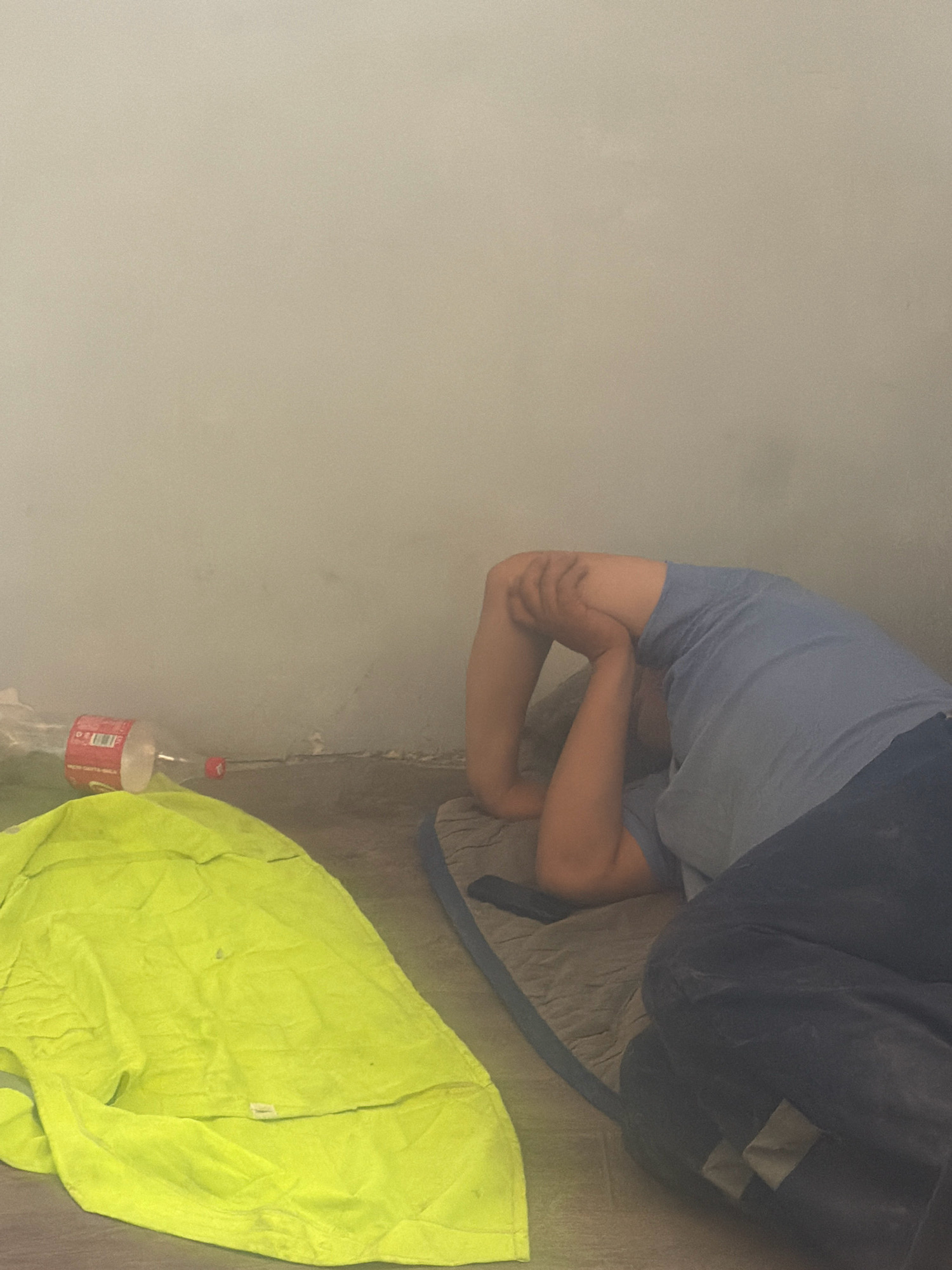 In Besapan, Uzbekistan, a person is lying on their back on a sleeping mat with their knees bent and hands clasped behind their head. The person is wearing a light blue t-shirt and dark blue pants. To the left of the person, there is a bright yellow garment, possibly a jacket or blanket, and a red and white soda bottle lying on its side. The environment appears to be indoors with a plain, light-colored wall and a floor that looks like concrete or a similar material. The overall impression is of a person resting or sleeping in a simple, perhaps temporary, living space.