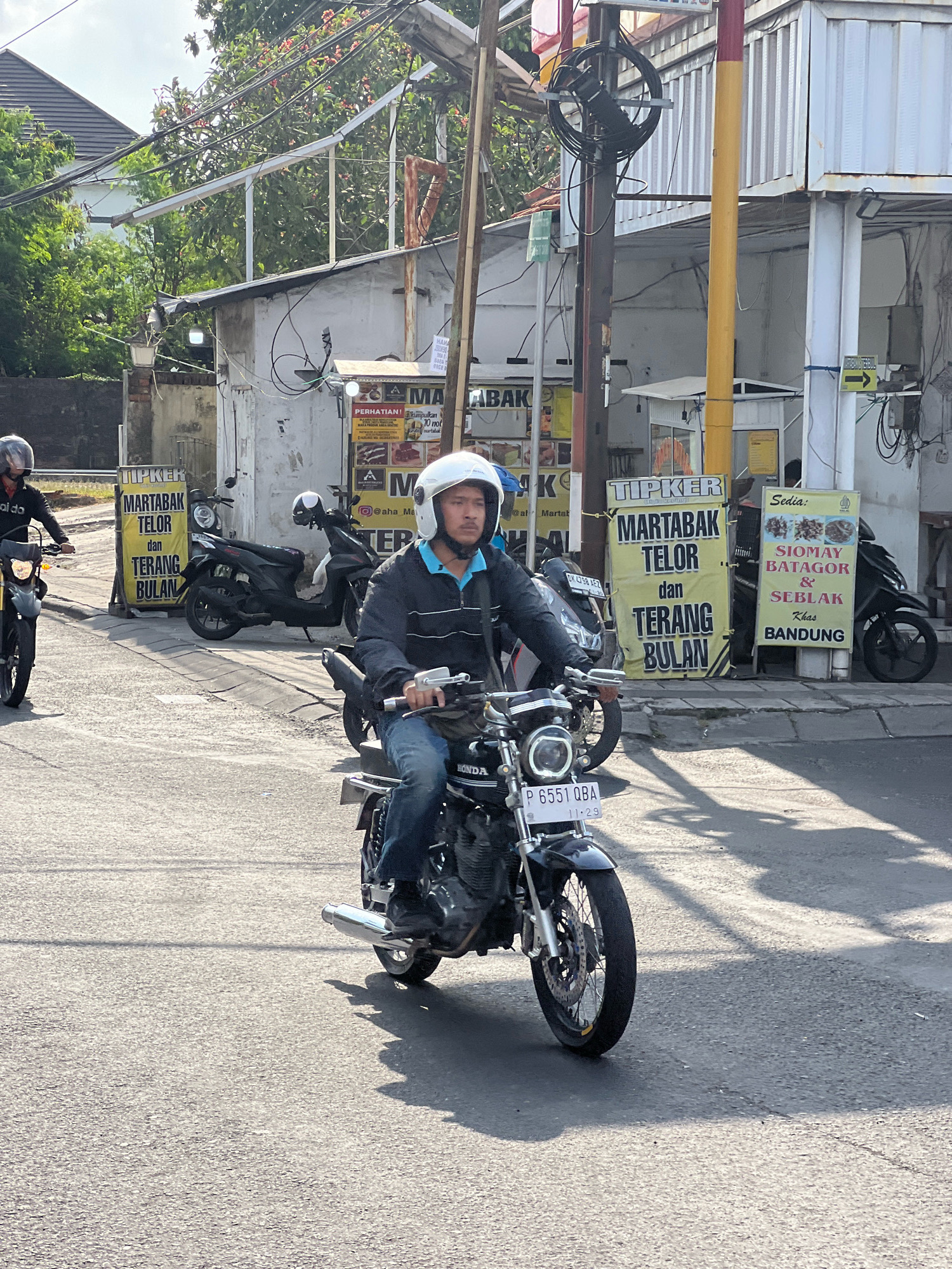 In Kuta, Indonesia, on a bright day, a man wearing a white helmet and blue shirt over a dark jacket is riding a black motorcycle. The motorcycle has a license plate that reads "P 6551 QBA". In the background, there are buildings and trees. A yellow sign on a pole reads "TIPKER MARTABAK TELOR dan TERANG BULAN". Another sign says "Sedia: SIOMAY BATAGOR & SEBLAK Khas BANDUNG". Two other motorcycles are parked on the side of the road, one black and one dark colored. The sky is cloudy and the sun casts shadows on the asphalt road.