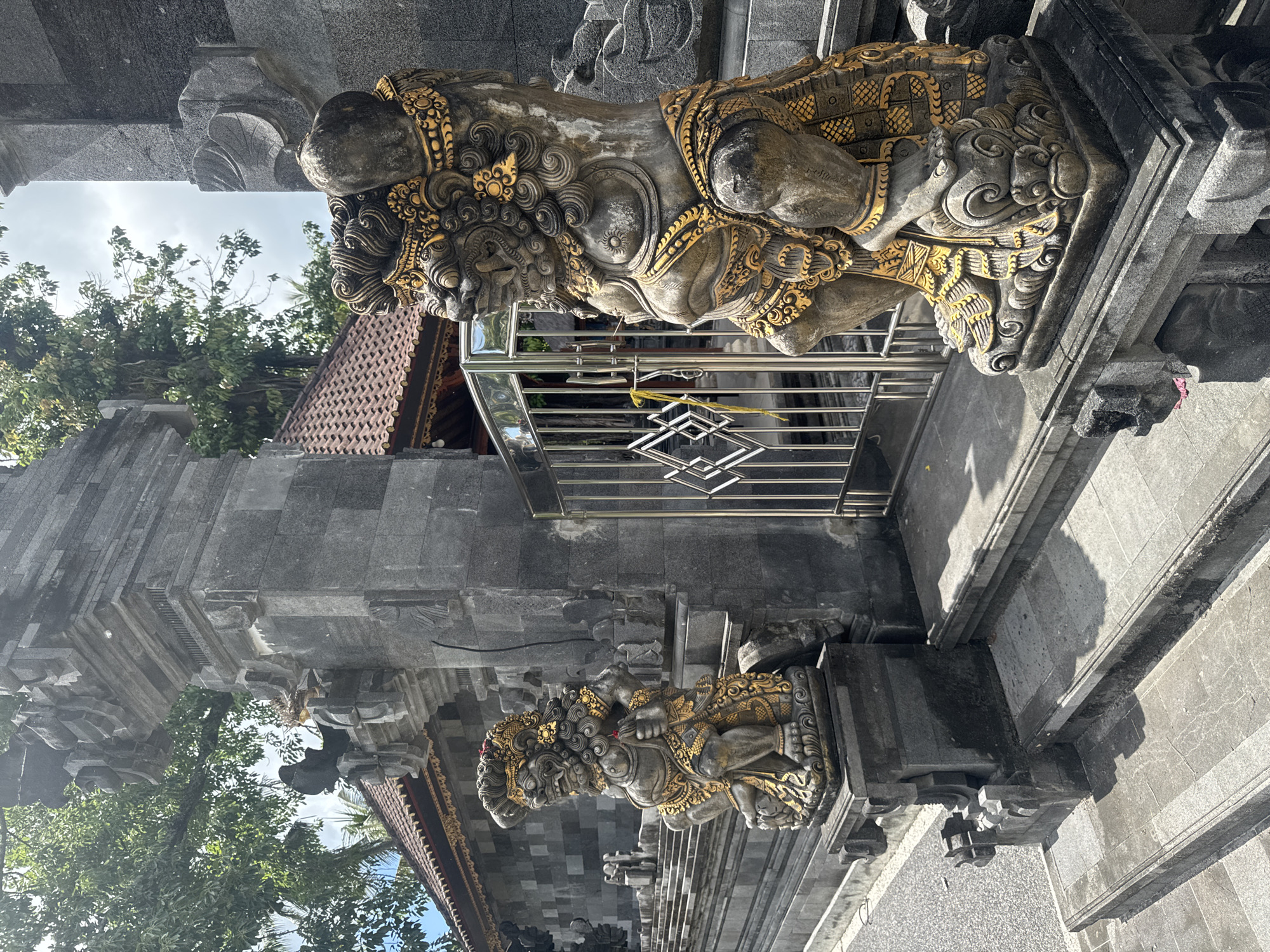 A stone temple entrance with two golden decorated statues on either side of a metal gate, located in Nusa Penida, Indonesia. The statues depict mythical creatures and are partially obscured by the gate. Trees and the temple roof can be seen in the background.