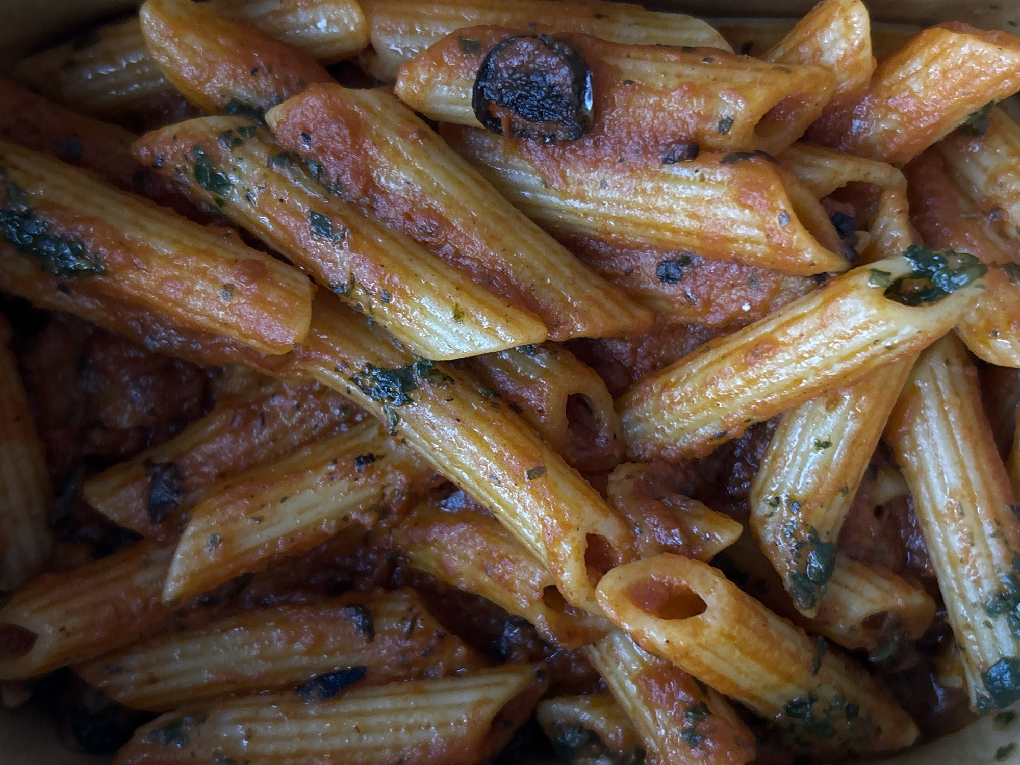 The image shows a close-up view of penne pasta coated in tomato sauce and garnished with what appears to be basil. A dark, circular object, possibly a cooked olive, is visible among the pasta. The setting is unspecified, and there's no information to suggest it's in Sofia, Bulgaria. There are no people visible. The environment suggests it could be a take-out container or a serving dish. The image is a still life, focused on the texture and color of the food.
