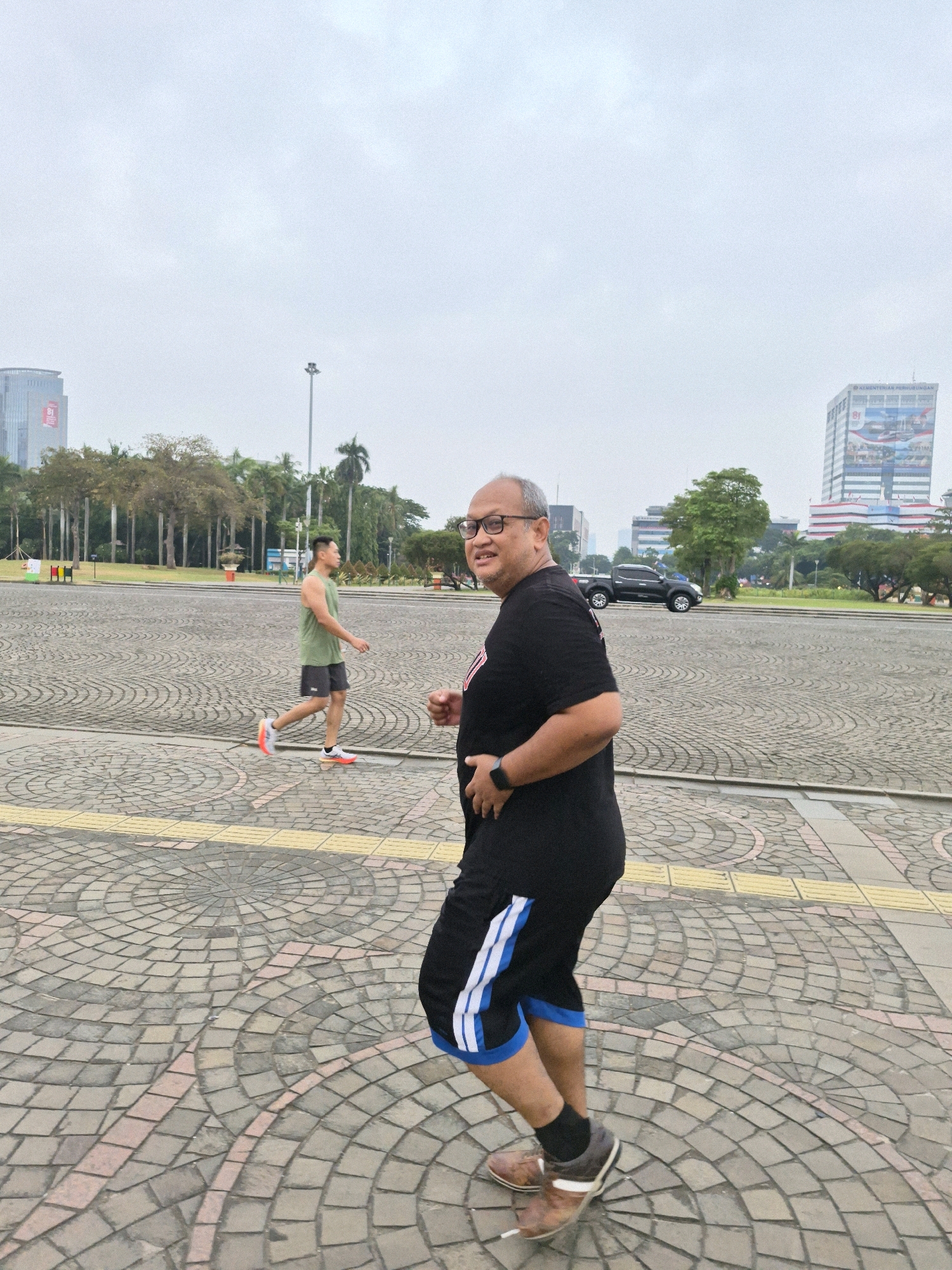 In the city of Central Jakarta, Indonesia, on an overcast day, a man in a black t-shirt and black shorts with blue and white stripes walks towards the camera on a patterned cobblestone surface. He is wearing glasses, a wristwatch, and brown shoes. To his left, another man in a green tank top and black shorts is jogging away from the camera. In the background, trees, buildings, a street with a black pickup truck, and a lamppost are visible.