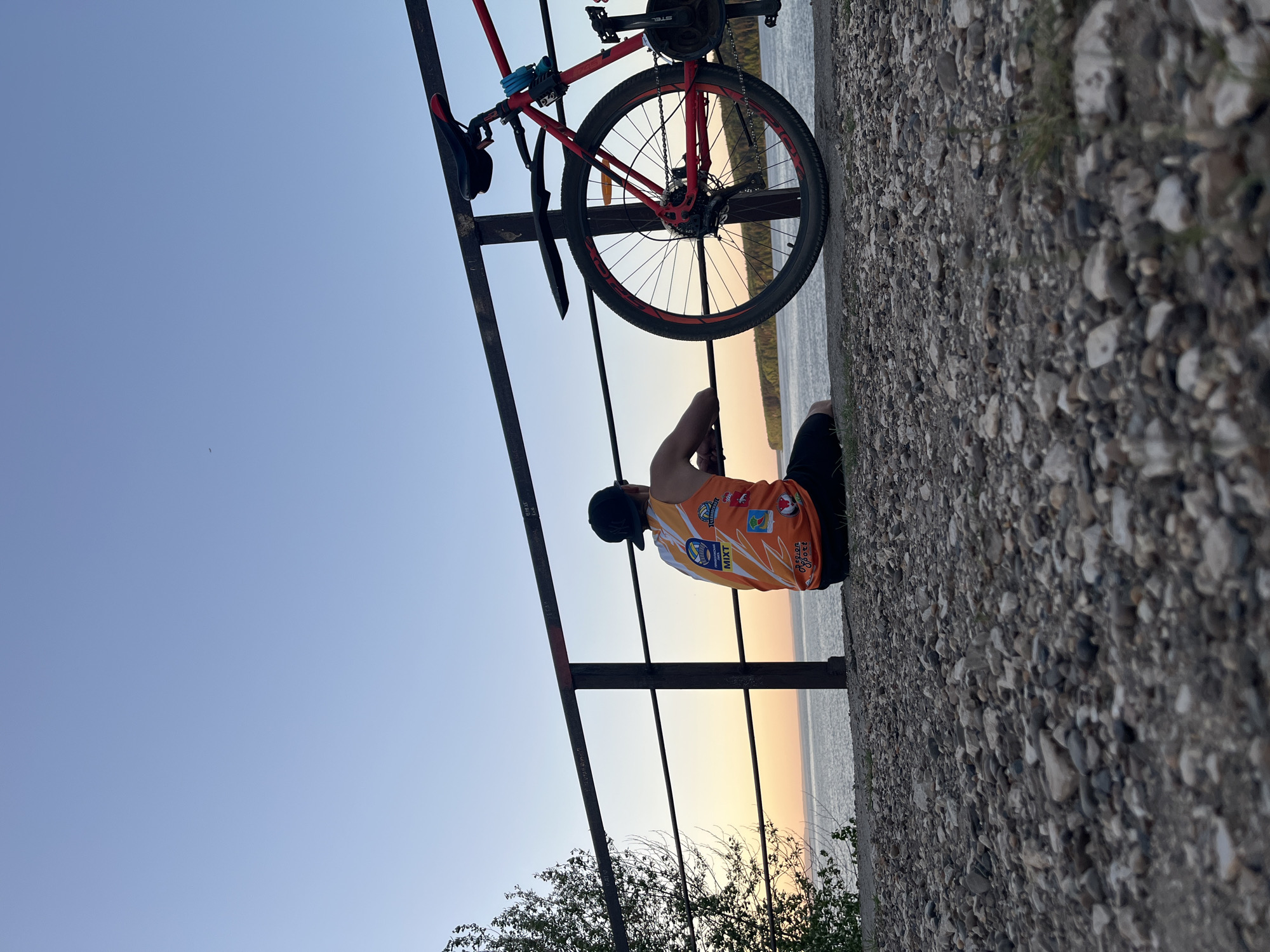 A man in a bright orange sports jersey and black shorts sits on a rocky ledge. He faces away from the viewer, looking out over a calm body of water. A bicycle, red and black, is secured to a metal railing next to him. The sky transitions from a light blue to a yellow-orange hue near the horizon. Some trees are visible in the lower left corner. The scene appears to be captured near the city of Chaikovsky, Russia, as suggested by text on the jersey.
