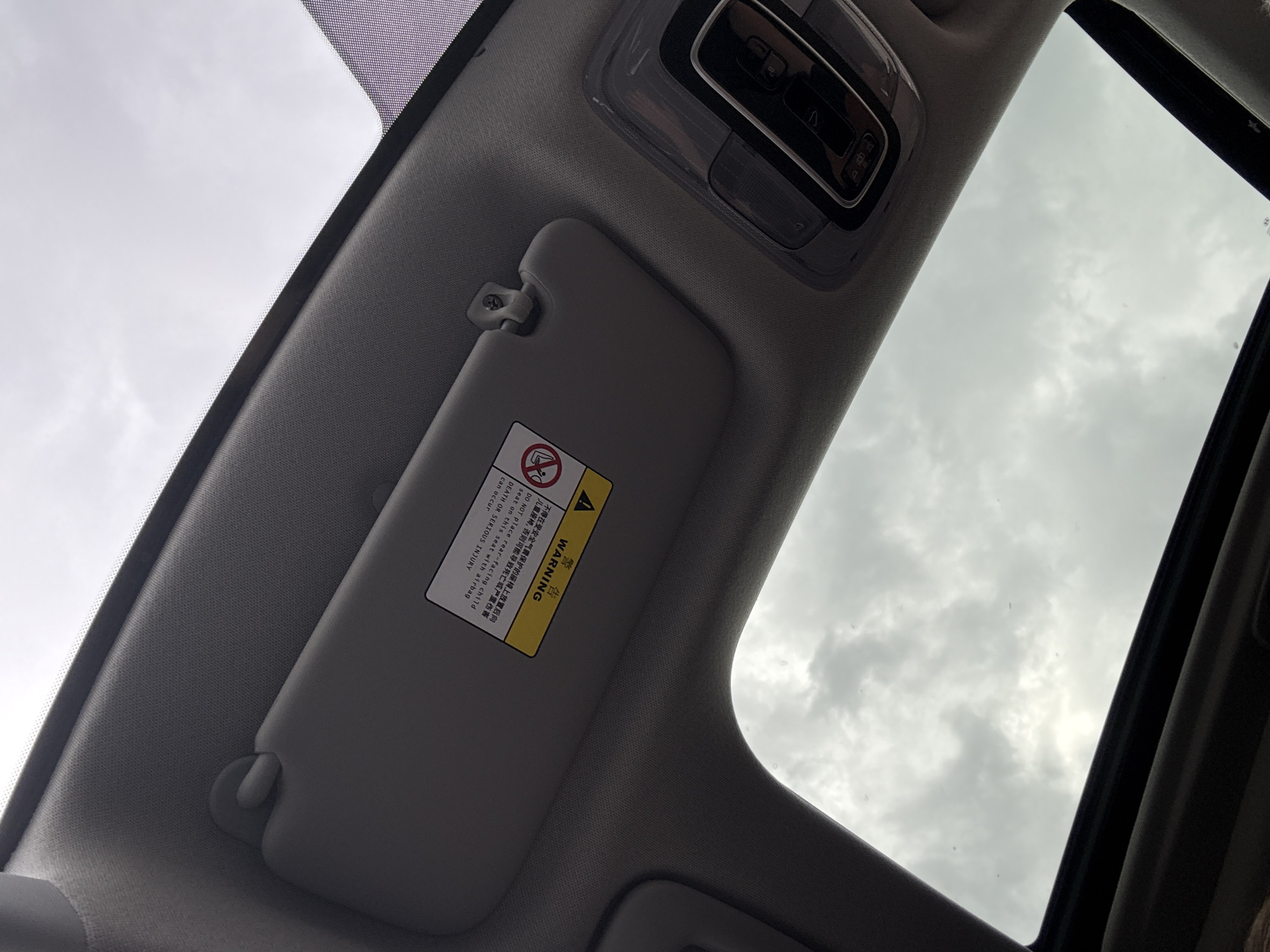 The image shows the interior of a vehicle, specifically the area around the sunroof and the driver's side sun visor. The sunroof is open, revealing a cloudy sky. The sun visor is folded down and has a warning sticker in both Chinese and English cautioning against placing a rear-facing child seat in a seat protected by an airbag. The car's interior ceiling light console is also visible.