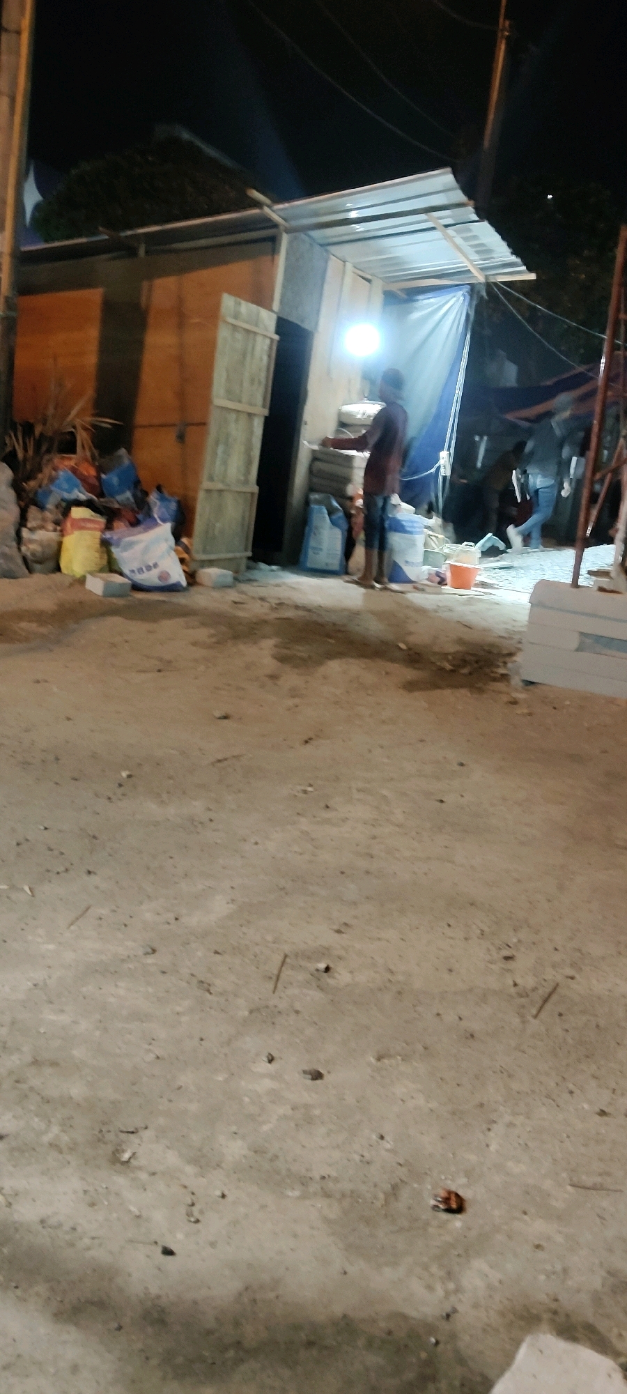 In Cibatu, Indonesia, at night, a person stands near a temporary structure with a corrugated metal roof. Bags of what appear to be cement or building materials are stacked on the left side of the scene. The ground is a mix of dirt and concrete. A bright light illuminates the area. In the background, other individuals are visible, suggesting ongoing activity. The overall scene depicts a construction or material storage area under artificial lighting.