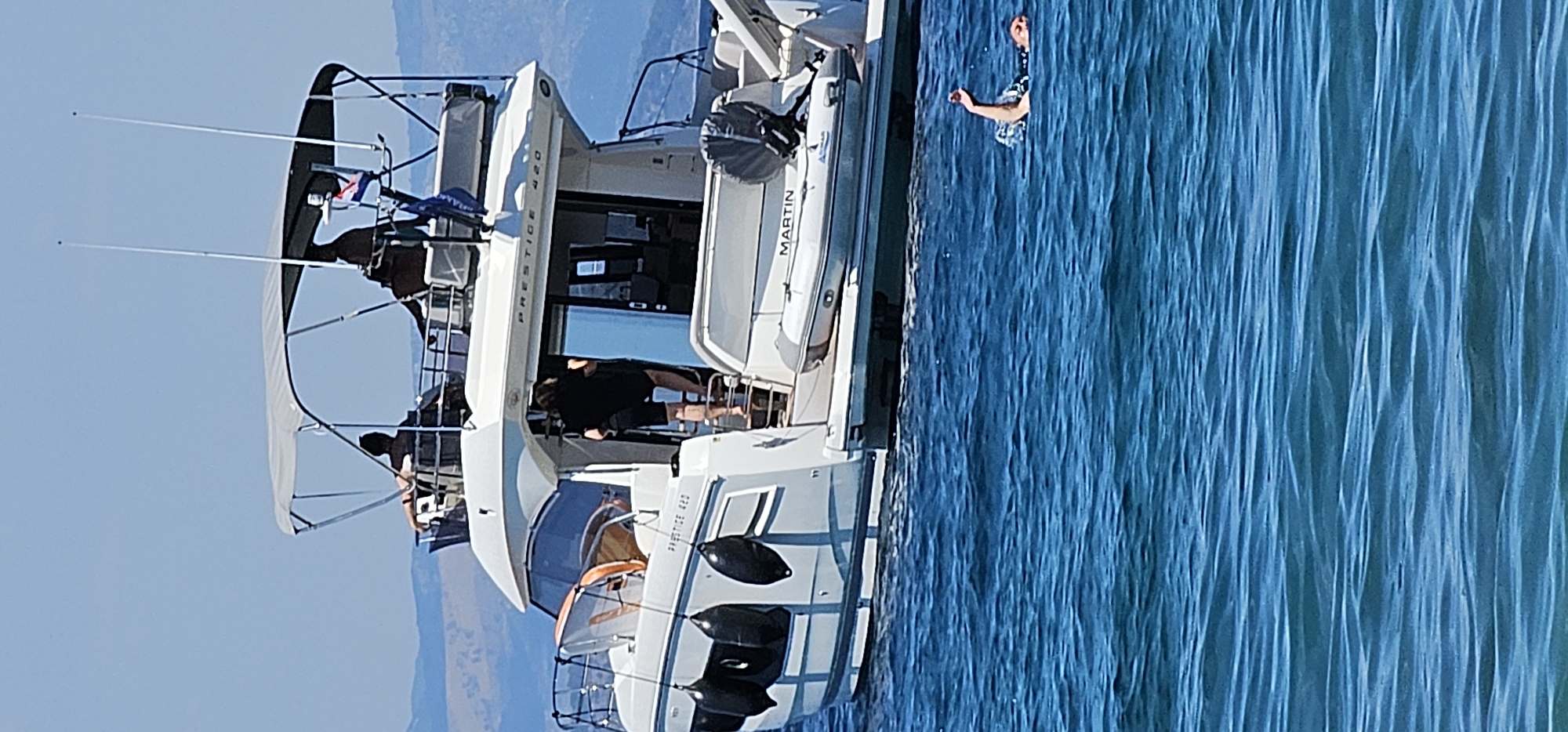 A person is swimming in the bright blue waters of the Adriatic Sea, off the coast of Supetar, Croatia. In the background, a large white motor yacht, identified by the name "PRESTIGE 420" and "MARTIN" visible on its hull, is anchored. The yacht has multiple levels and a Bimini top providing shade. Several people are visible on the deck of the yacht, some seated and others standing. One person is seen descending stairs from the upper deck to a lower deck. The water is choppy, with visible waves. The sky is clear and blue, with a hazy mountainous coastline in the distance.