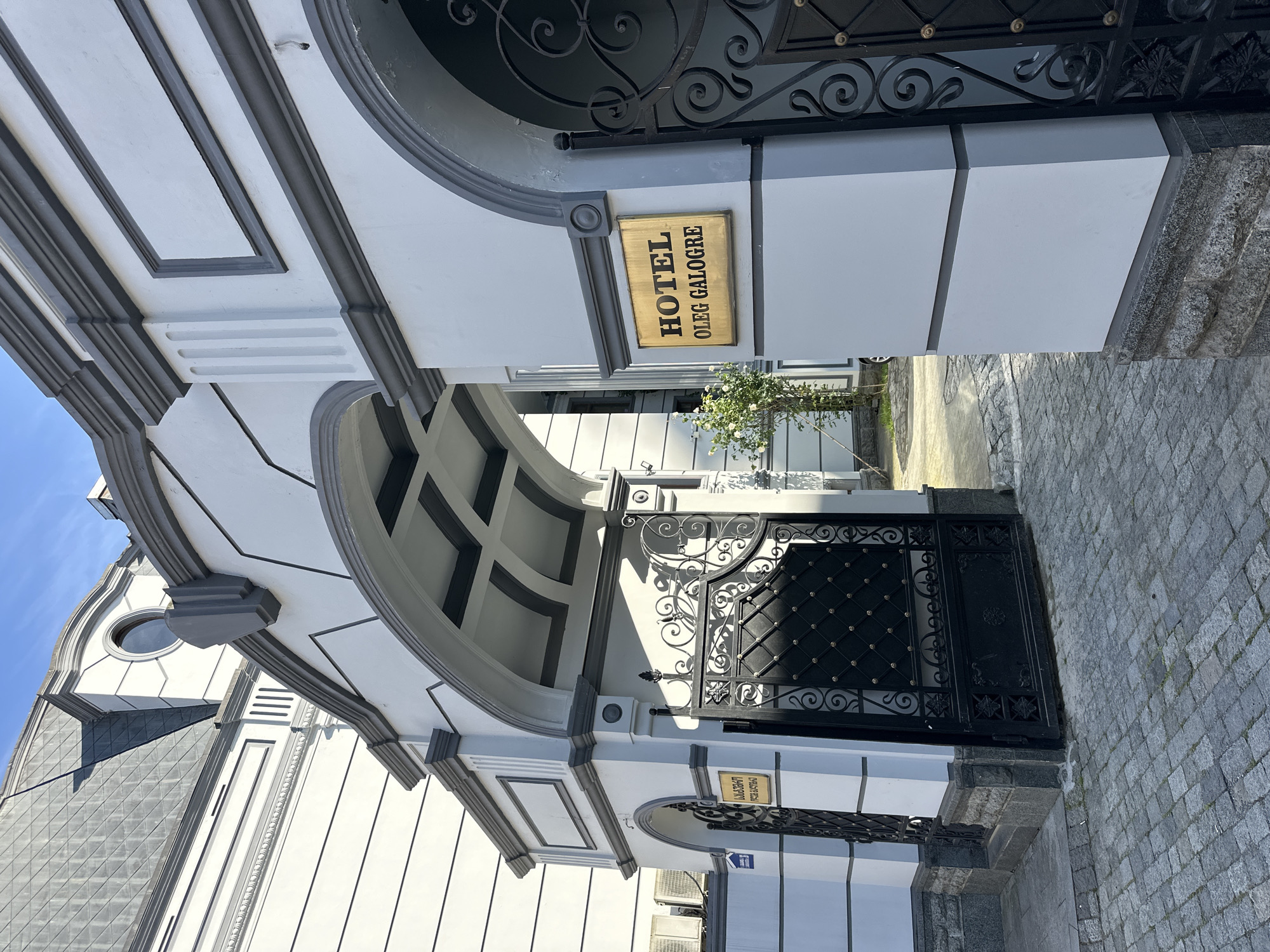 The image shows the entrance to "Hotel Oleg Galogre" in Batumi, Georgia. The entrance features a white and grey arched gateway with a decorative black iron gate. A gold-colored sign displays the hotel's name in vertical orientation. The building's facade is white with grey trim. Pavement stones are visible in the foreground. There are no people in the shot. A small tree is visible in the background, partially obscured by the gate.