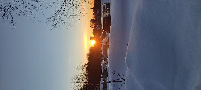 A winter sunset paints the sky in hues of orange and blue, backlighting distant buildings and bare trees. Cars parked along a snow-covered street glisten with reflected light. A thick blanket of fresh snow dominates the foreground. Silhouetted branches reach into the sky from the top left.