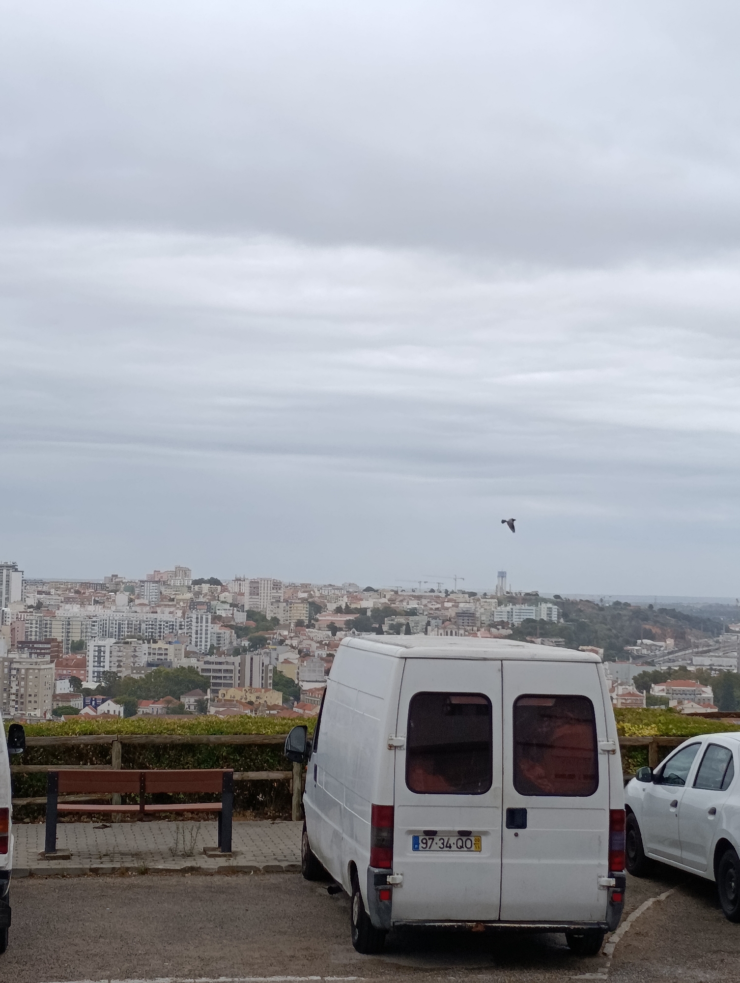 The image shows a cloudy day with a view of the city of Setúbal, Portugal, from an elevated position. In the foreground, there are two white vans parked on a paved area. One van, a larger cargo van, is positioned in the center of the frame, facing away from the camera. It has a license plate that reads "97-34-QQ". To the right of this van, a white sedan is partially visible. A wooden bench with a slatted seat is situated behind the vans, separated by a paved walkway. A wooden fence runs along the edge of the paved area, behind which is a dense hedge. Beyond the hedge, the city of Setúbal stretches out, a densely packed landscape of buildings with varying roof colors. In the sky, a single bird is in flight. The overall impression is one of urban sprawl under an overcast sky.