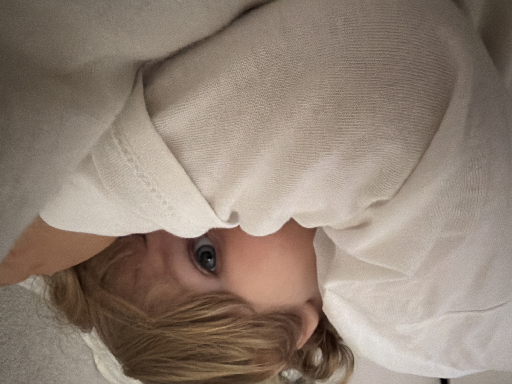 A young child with blonde hair and blue eyes is partially covered in a white cloth or blanket. The child's face is visible, peeking out from beneath the fabric. The photo appears to be taken indoors. The location and specific actions and interactions are not discernible.