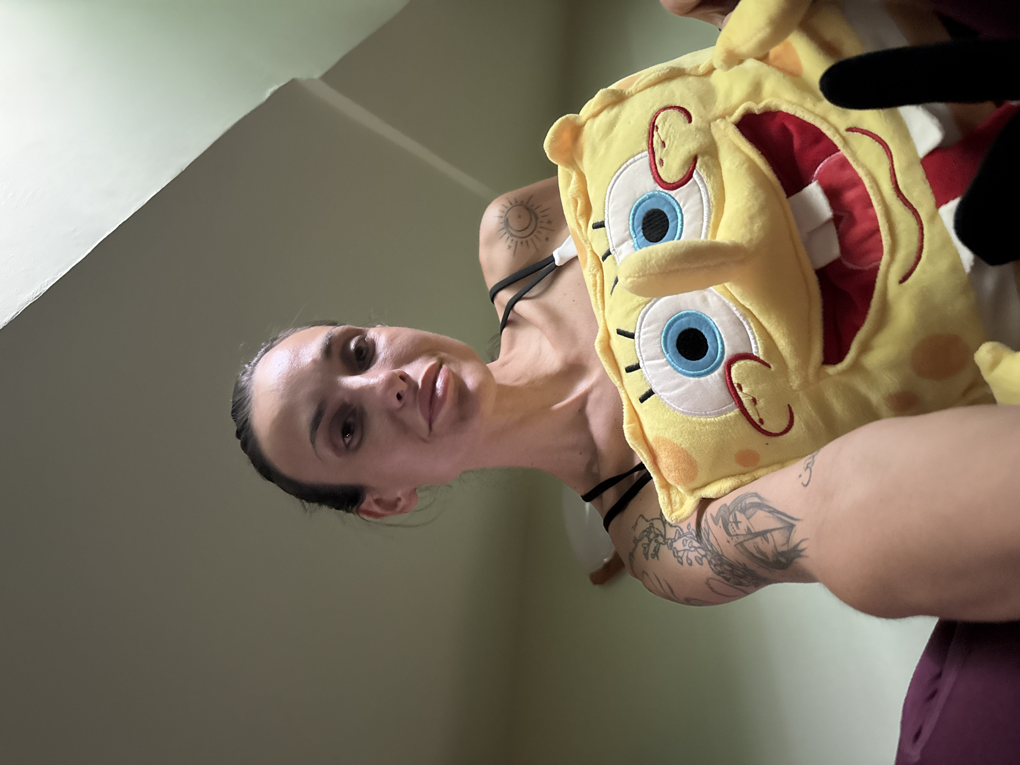 A low-angle, close-up portrait, rotated counter-clockwise, depicts a person holding a unique plush toy. The individual, likely an adult female, is positioned centrally, looking directly at the camera with a neutral expression. Her dark hair is pulled back from her face. She wears a black top with thin straps. Visible tattoos adorn her arms and shoulder; one on her upper left arm features a sun and crescent moon design, while other abstract patterns are seen on her lower arms.
She is holding a bright yellow plush toy shaped like the cartoon character SpongeBob SquarePants. The toy has prominent blue eyes, red accents around the cheeks and mouth, and is distinctive for an opening on its chest that reveals red fabric resembling internal organs, suggesting an "anatomical" or "guts out" design. A dark-colored object, possibly another plush or a hand, is partially visible near the top right of the SpongeBob toy.
The background consists of plain, light-colored walls, featuring a visible corner where two walls meet in the upper left of the frame. The lighting is soft and diffused, characteristic of indoor ambient light. The scene portrays a casual, indoor moment, possibly within a residence in Pleven, Bulgaria. There is no visible text in the image. The person's direct gaze indicates they are posing for the photograph.