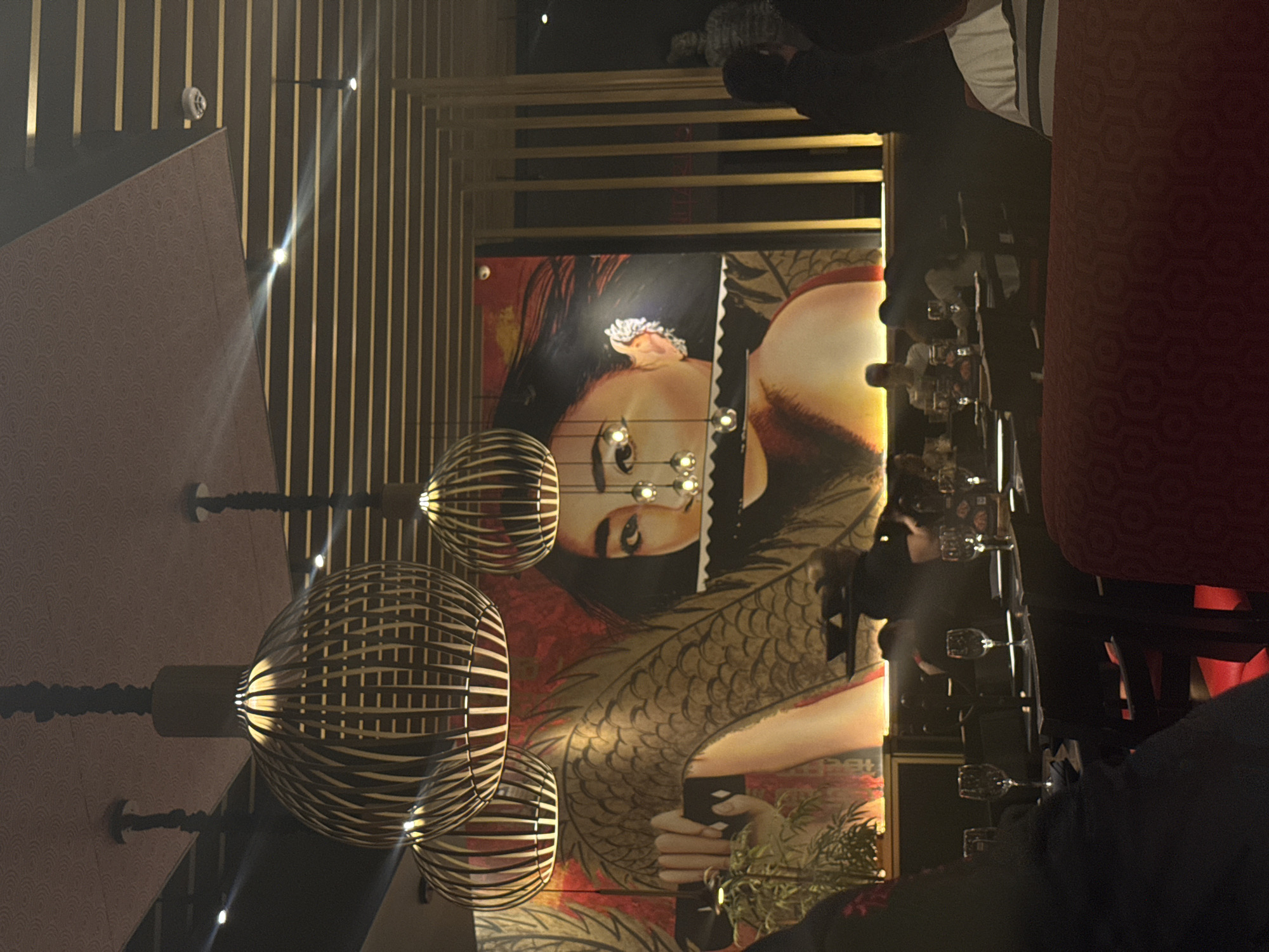 This image shows the interior of a restaurant in Mexico City, Mexico. The ceiling is adorned with hanging, spherical, wooden-slatted lamps. A mural of a woman with a dragon motif is visible on the wall. Tables are set with glassware, and people are seated, some partially visible in the image. The interior design features vertical gold stripes along the walls.