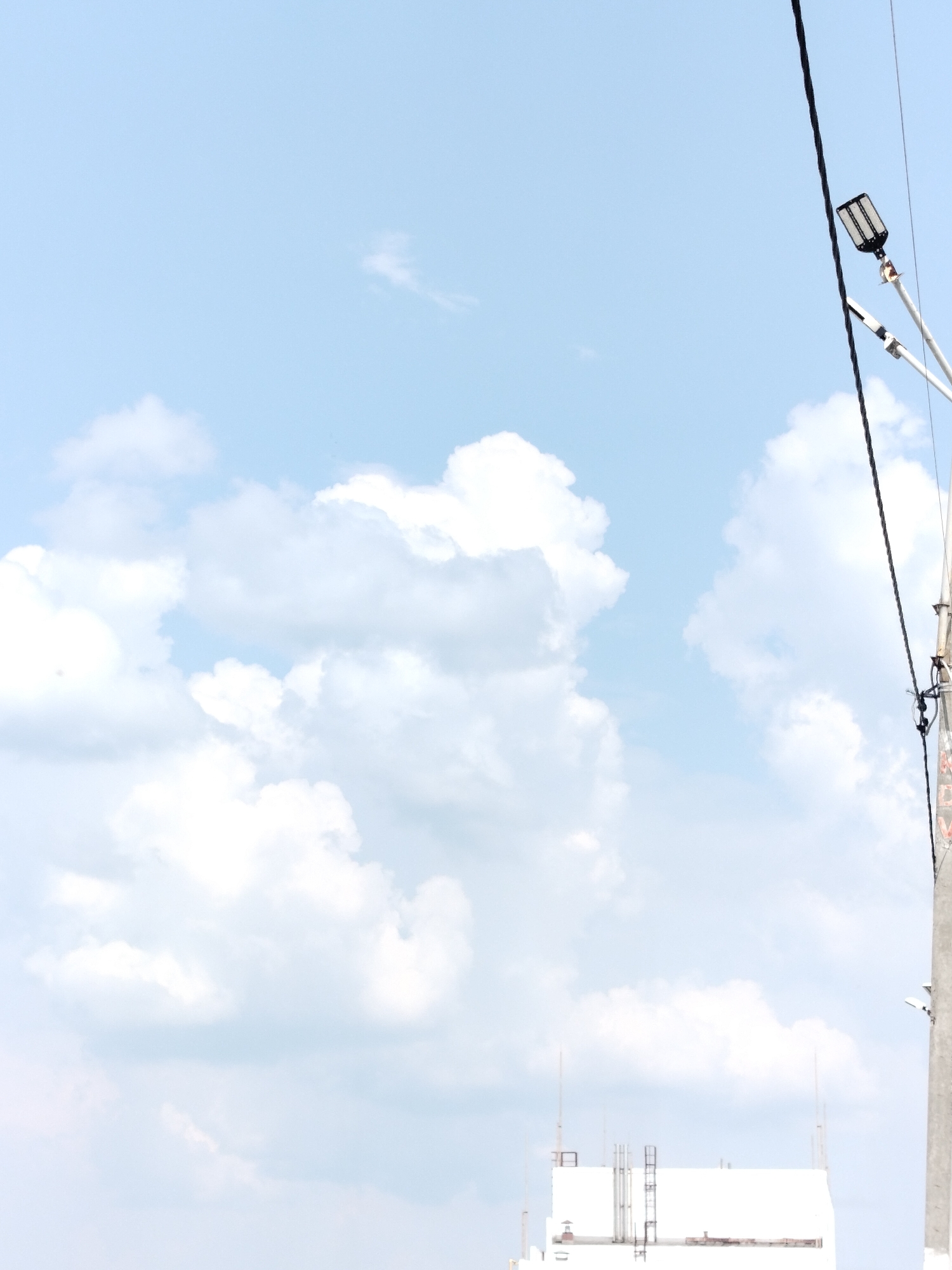 The image shows a bright, overcast sky with large, fluffy white clouds. In the foreground, a power pole with a black cable and a modern-looking streetlamp is visible on the right side of the frame. The streetlamp is angled slightly downwards and to the left. At the bottom of the image, a white building with some visible infrastructure, including what appear to be antennas or railings, is partially visible. There are no people or animals in the frame. The overall scene suggests an outdoor urban or semi-urban environment under a daytime sky. The location in Yashkino, Russia, is not visually identifiable from the image alone.
