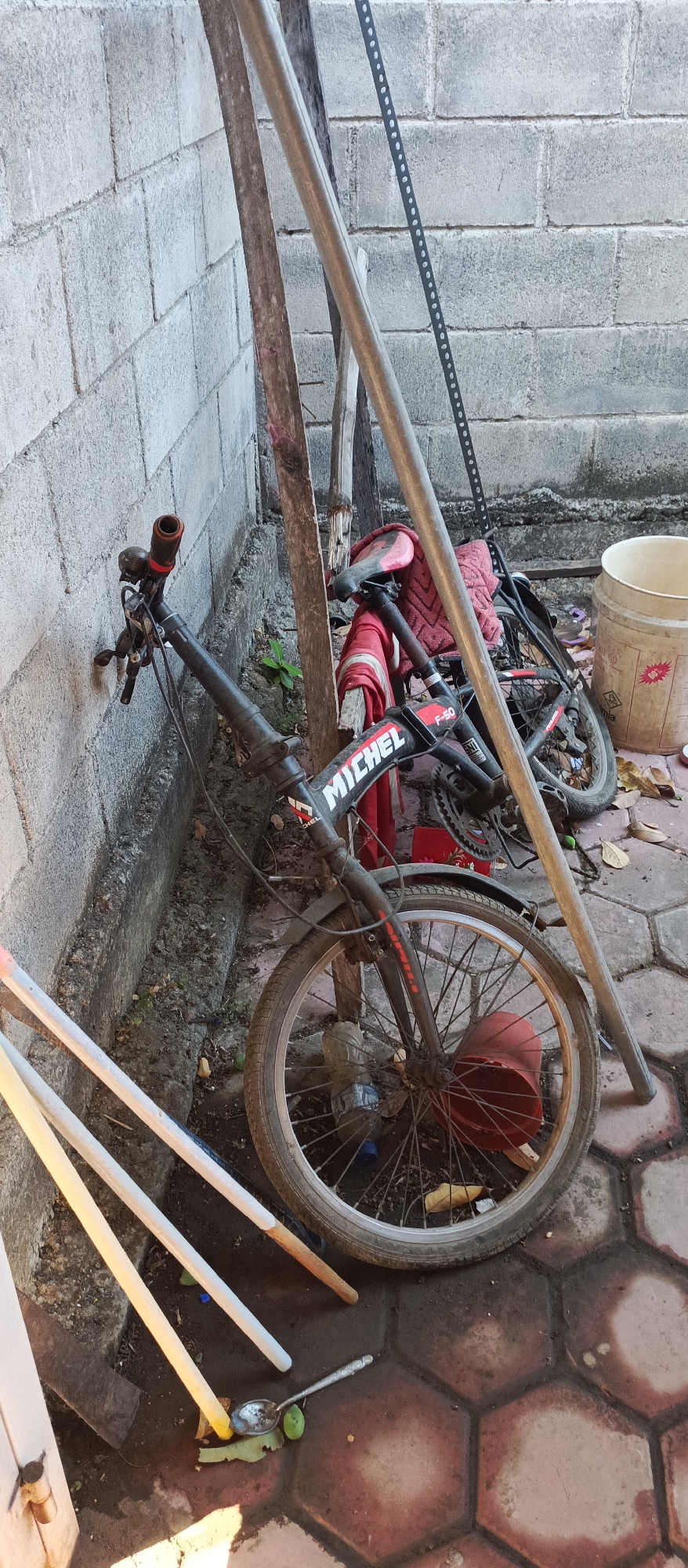 An exterior, medium shot captures a black and red bicycle leaning against a gray cinder block wall. The bicycle is positioned at an angle, with its front wheel in the foreground. The handlebars are turned to the left, and a red cloth is draped over the seat and part of the frame. The word "MICHEL" is visible on the bicycle's frame. To the left of the bicycle, three long, pale poles are partially visible, angled towards the ground. A metal pole runs vertically behind the bicycle, and a perforated metal strip is attached to the wall on the right side. The ground is paved with hexagonal tiles. A light-colored plastic bucket with a red sticker is situated to the right of the bicycle. Fallen leaves and debris are scattered on the ground. The scene appears to be an outdoor storage area or alleyway.