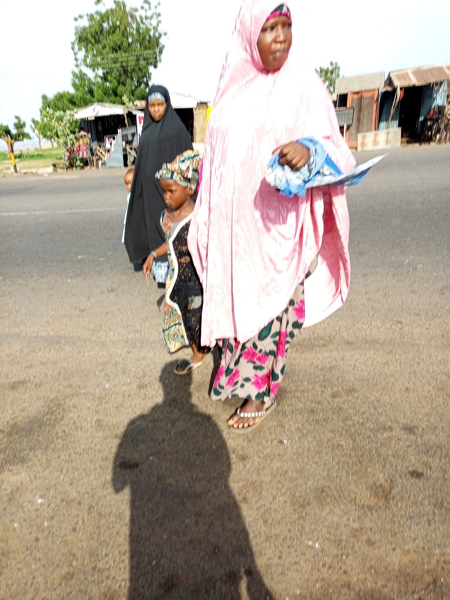 The image shows three people, two women and a child, walking on a paved road in Gadao, Nigeria. The woman in the foreground is wearing a light pink hijab and a long floral dress. She is holding a blue and white patterned fabric in her right hand. To her left, a child in a patterned dress and headscarf walks ahead. Behind them, a woman in a black hijab is also visible. The scene appears to be set outdoors during daylight, with a building resembling a small shop or stall in the background. The ground is paved, and a shadow of the people is cast on the road. The overall mood seems ordinary, suggesting a daily activity. No visible text is present in the image.